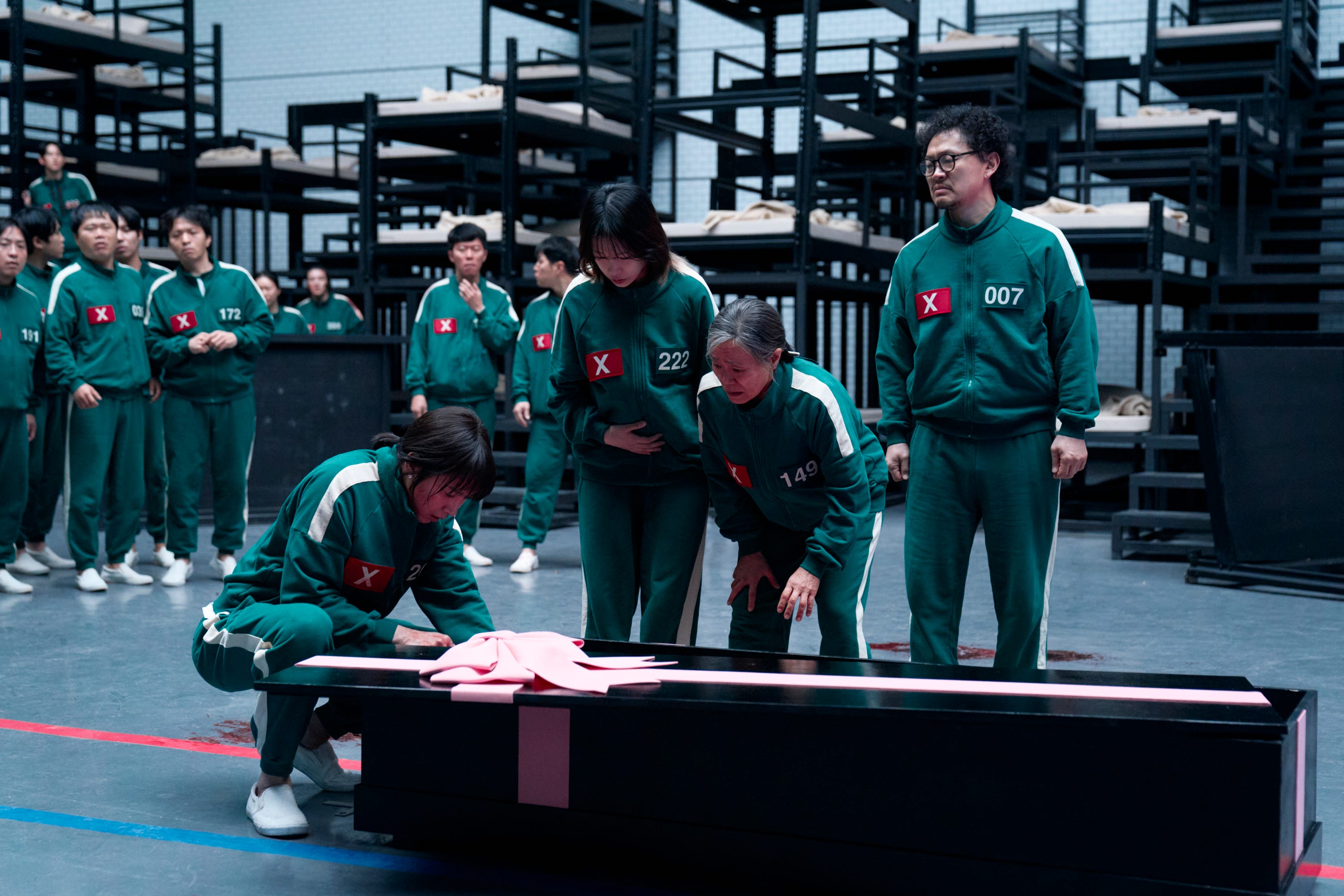 A still of "Squid Game" showing Park Sung-hoon, Jo Yu-ri, Kang Ae-sim and Yang Dong-geun wearing green tracksuit, standing around a coffin with a pink bow on it.