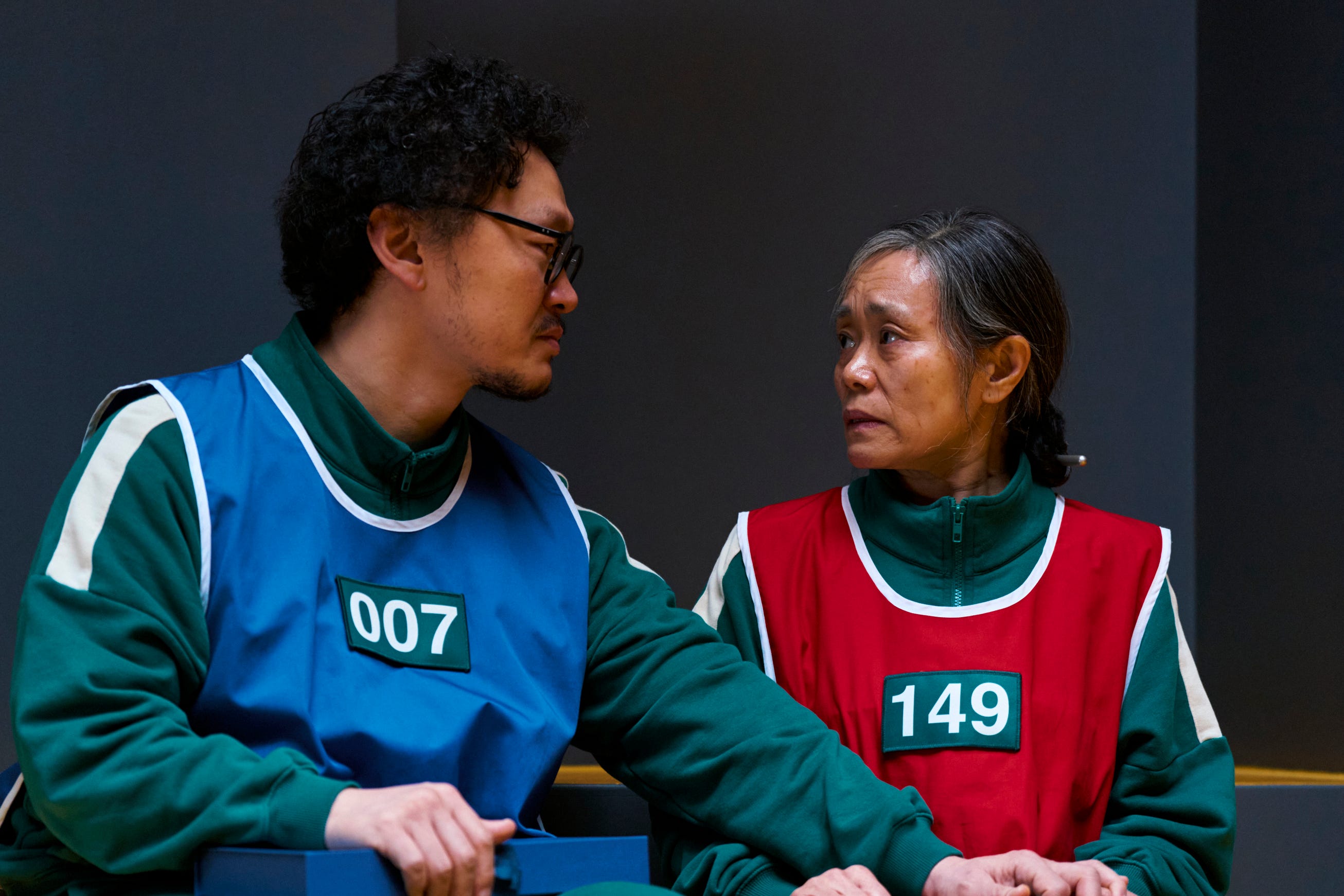 A still of "Squid Game" season 3 showing Yang Dong-geun wearing a blue vest over a green jumpsuit and Kang Ae-sim wearing a red vest over a green jumpsuit.