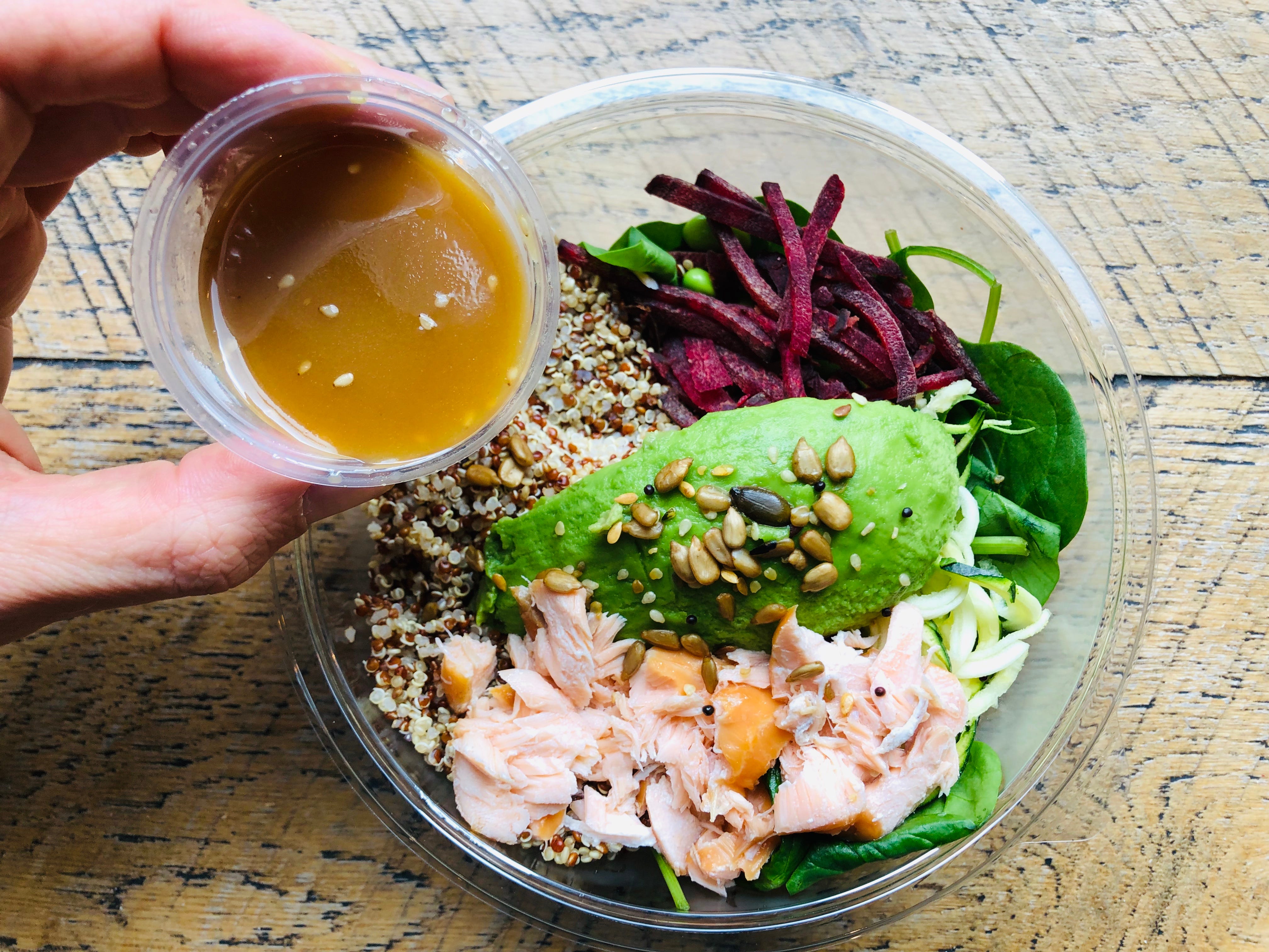 Cup of dressing being poured over beets, avocado, seeds, and pulled chicken in bowl