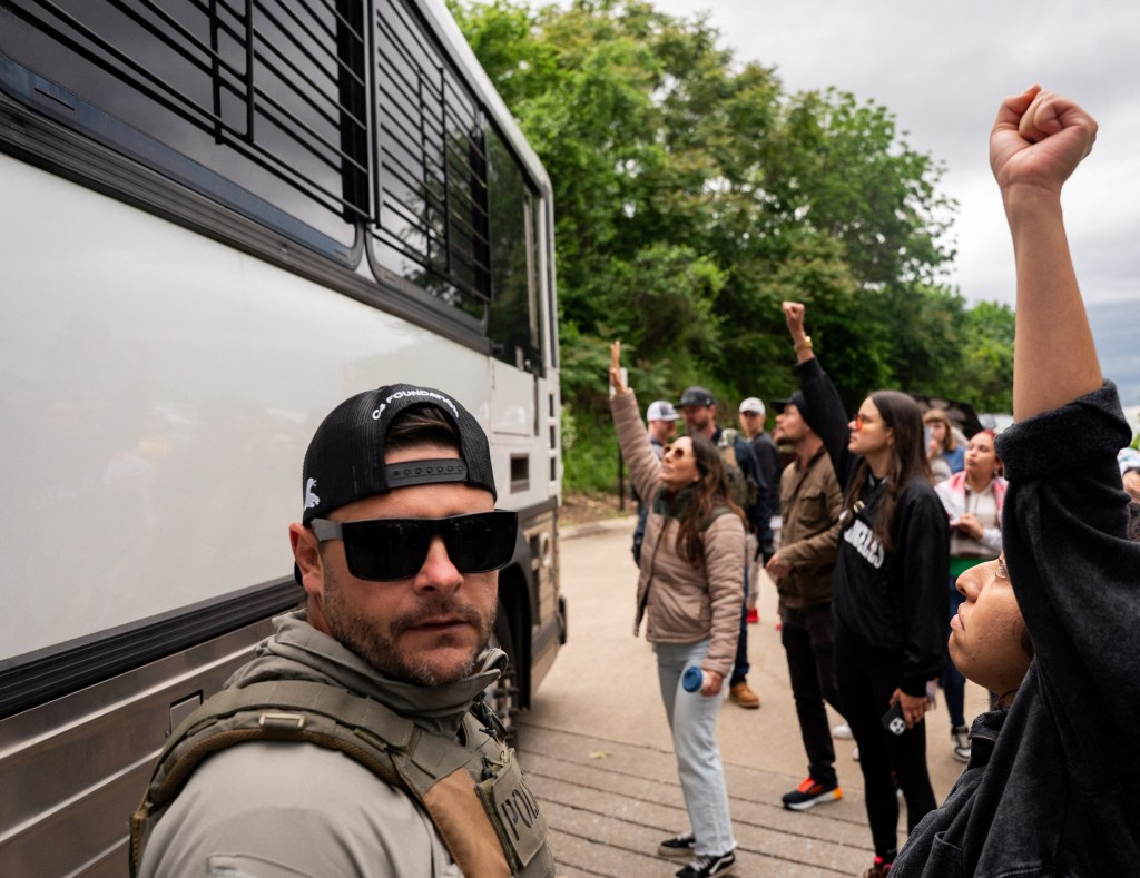 Protesters raise their arms towards a bus carrying detained individuals from the field office.