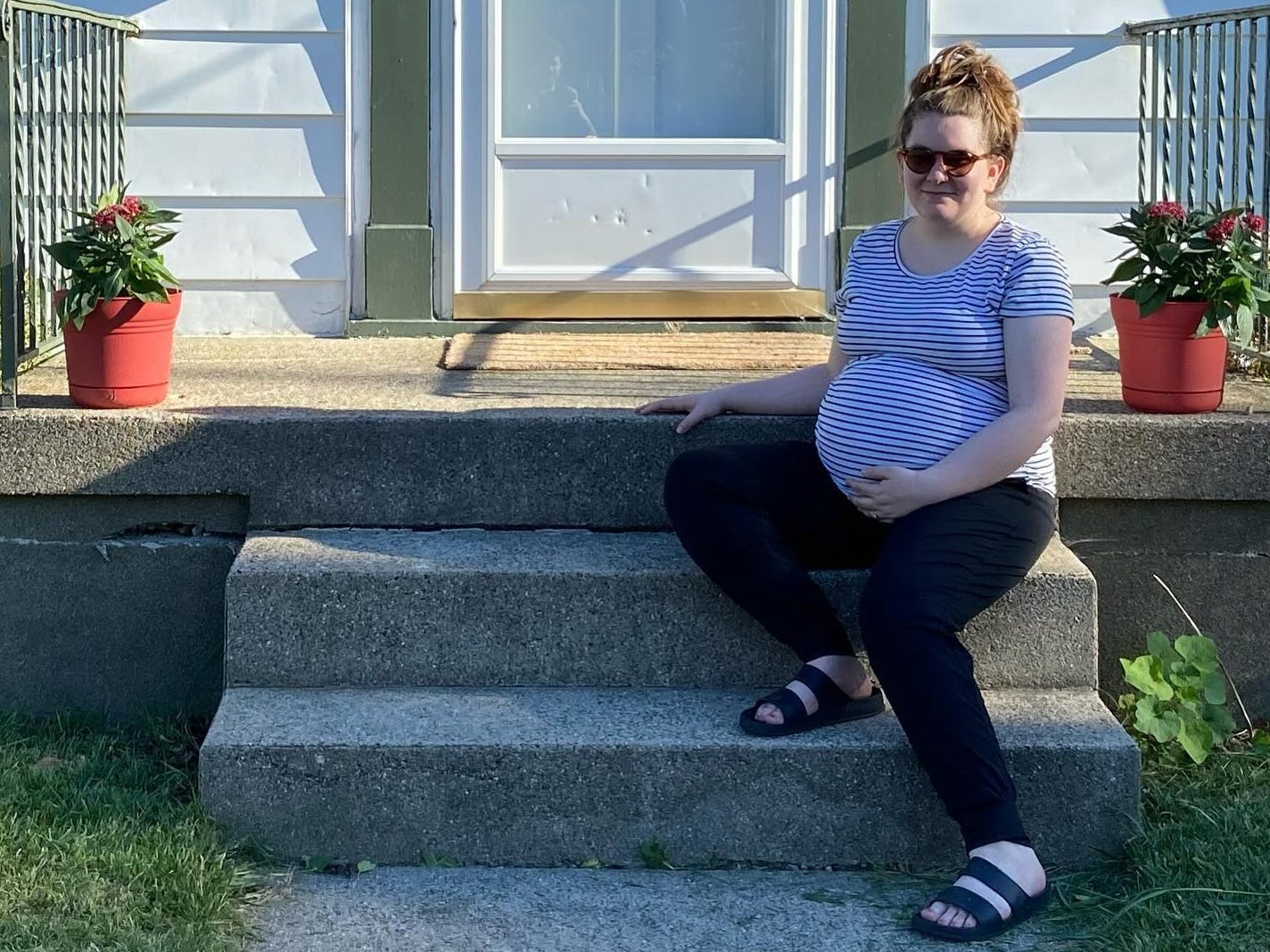 The author is pregnant and sitting on steps while wearing sunglasses, a t-shirt, and black pants, and holding her belly.