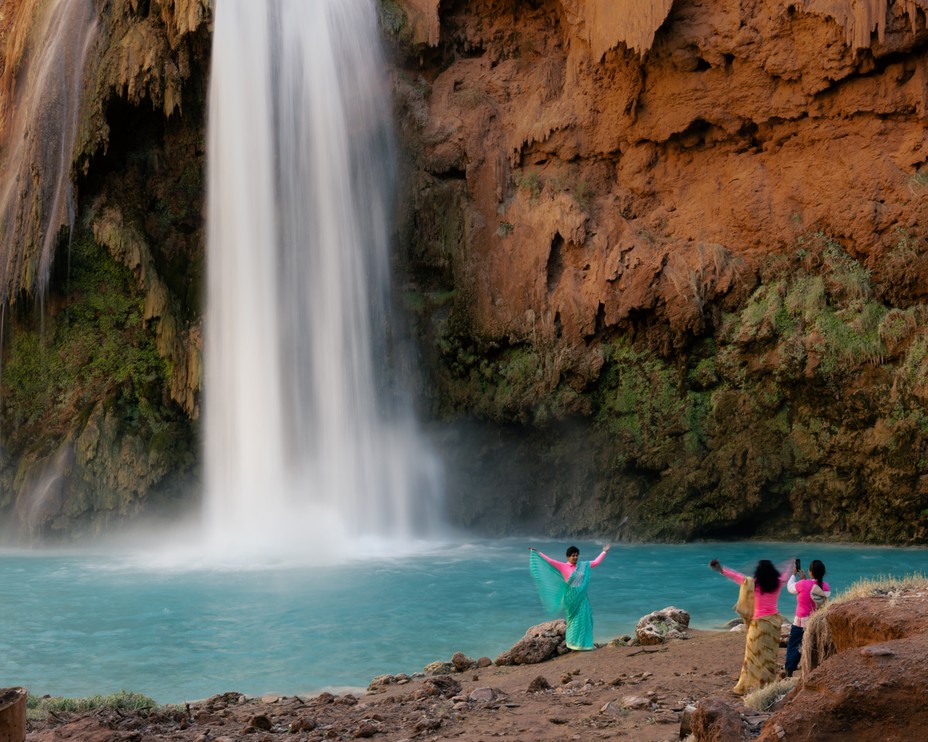 photo of women in brightly colored clothing taking photos with outstretched arms in front of large waterfall and pale-blue pool