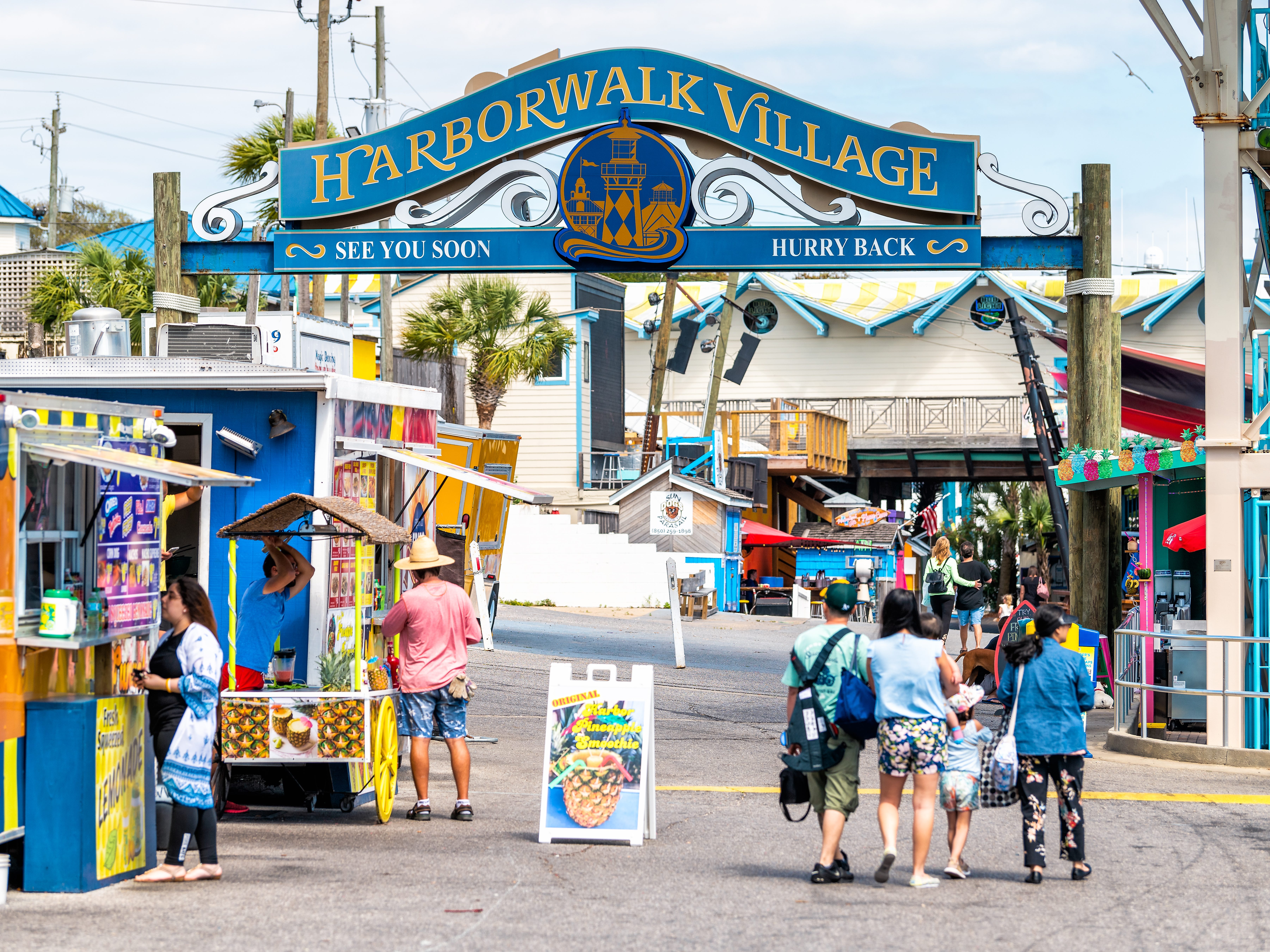 People walking around a shopping area with food trucks and a sign that reads, "Harborwalk Village."