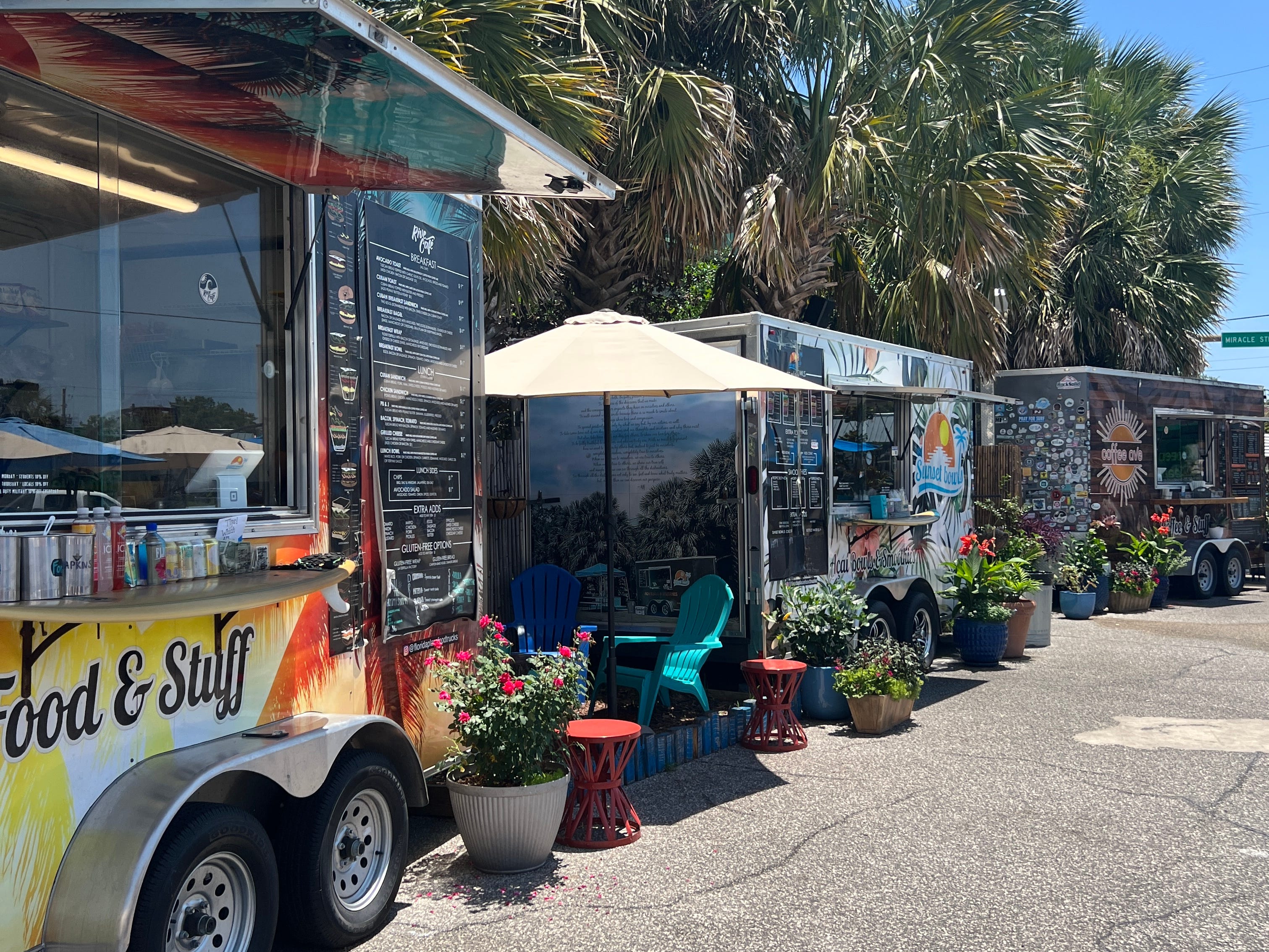 Three food trucks parked along the side of a road, with palm trees behind them.