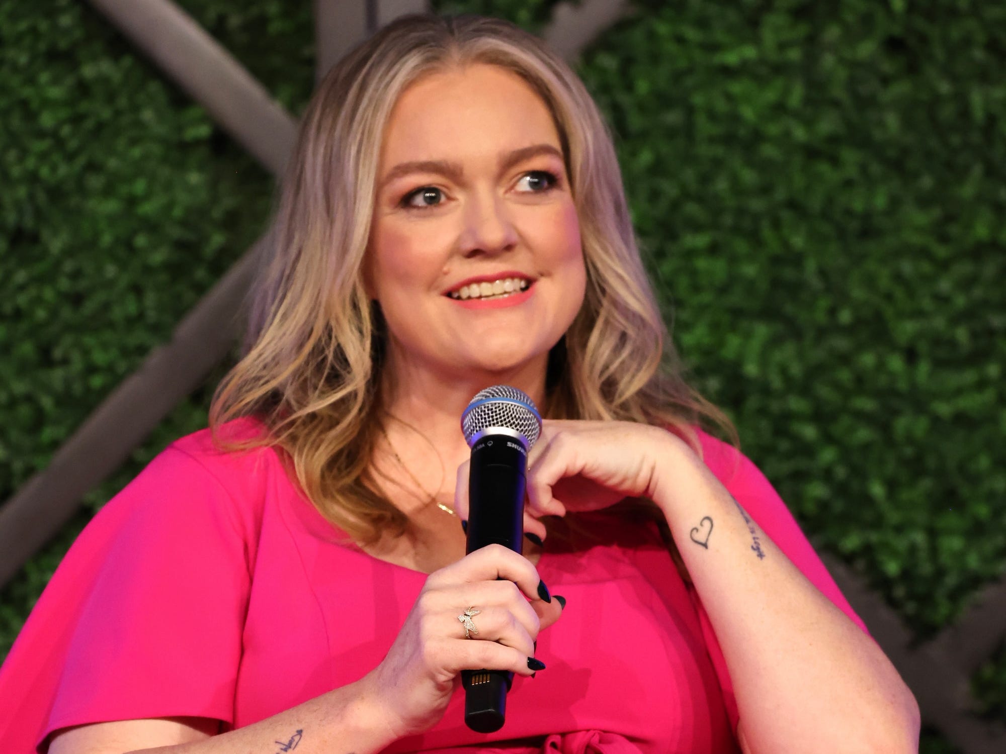 Colleen Hoover sits in a chair in front of a green wall, holding a microphone.