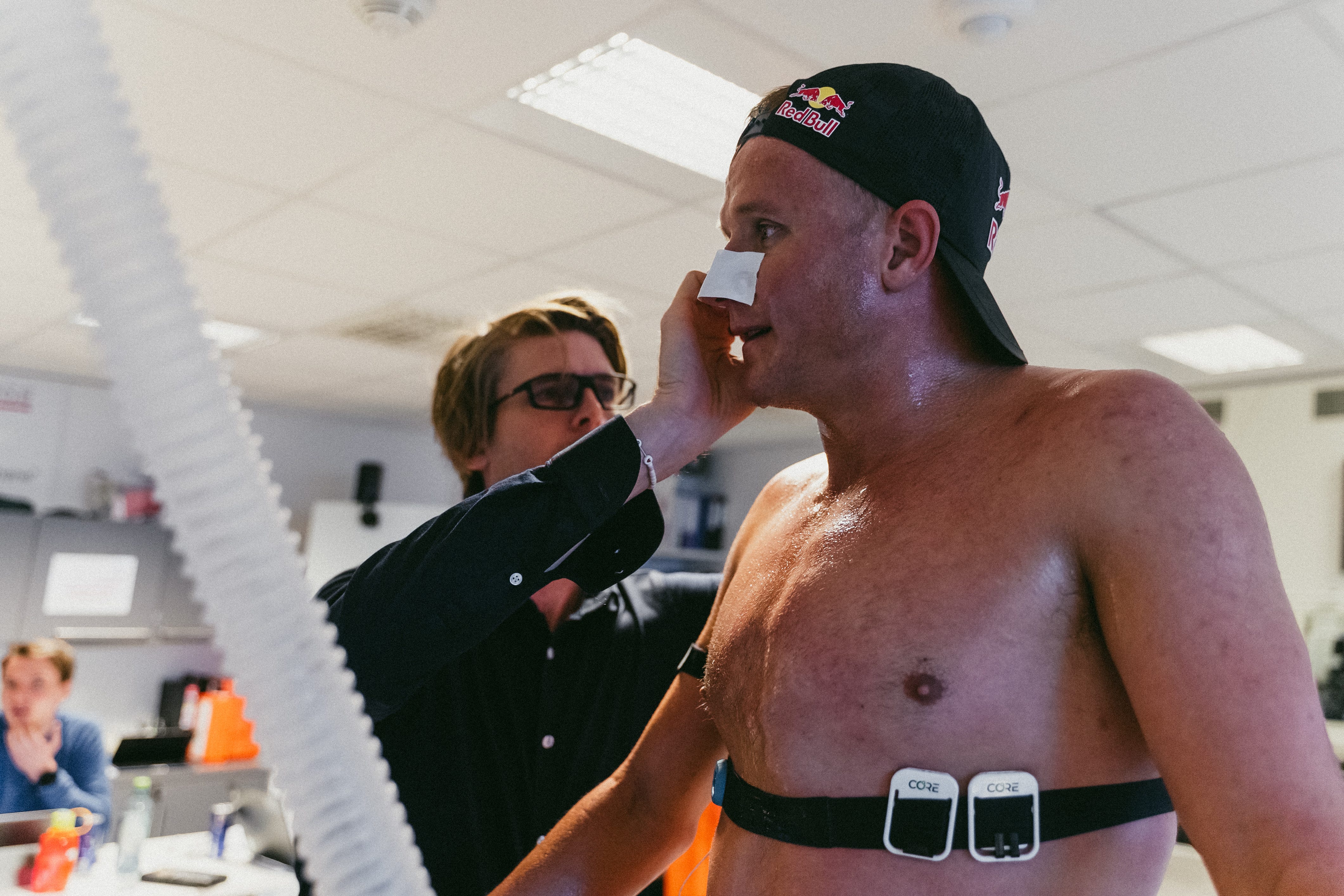 A shirtless athlete in an exercise science lab is wearing CORE heat sensors, assisted by his coach.