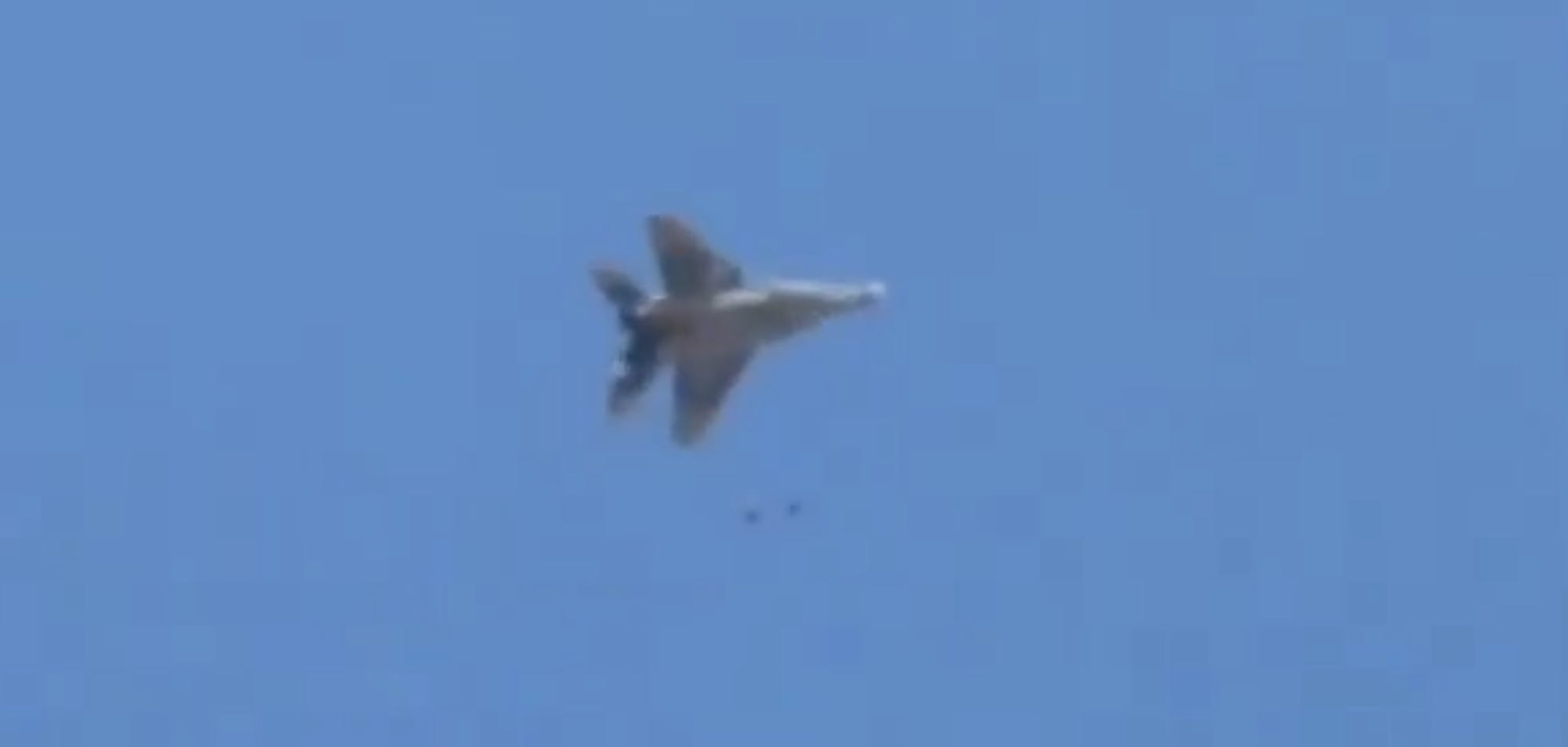 A video still shows a grey jet against a blue sky with a smaller item falling from it
