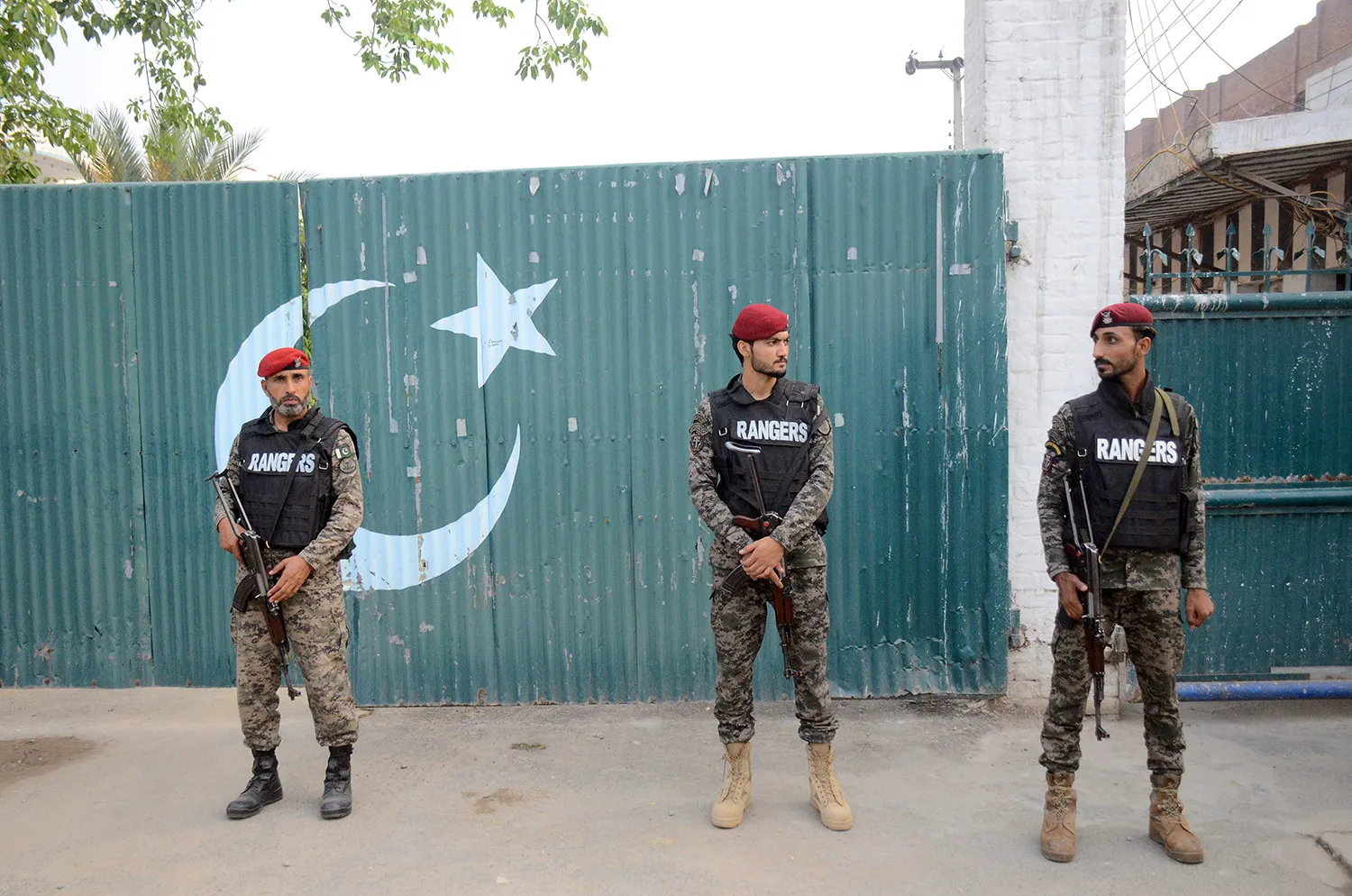 Three soldiers with guns and wearing fatigues and berets stand in front of a metal barrier with the flag of Pakistan painted on it.