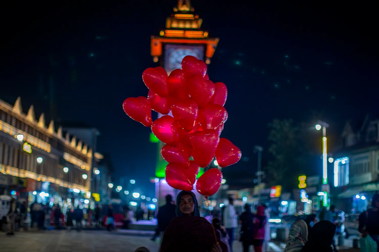 A woman holds red heart balloons in a night scene in front of an illuminated clock tower.