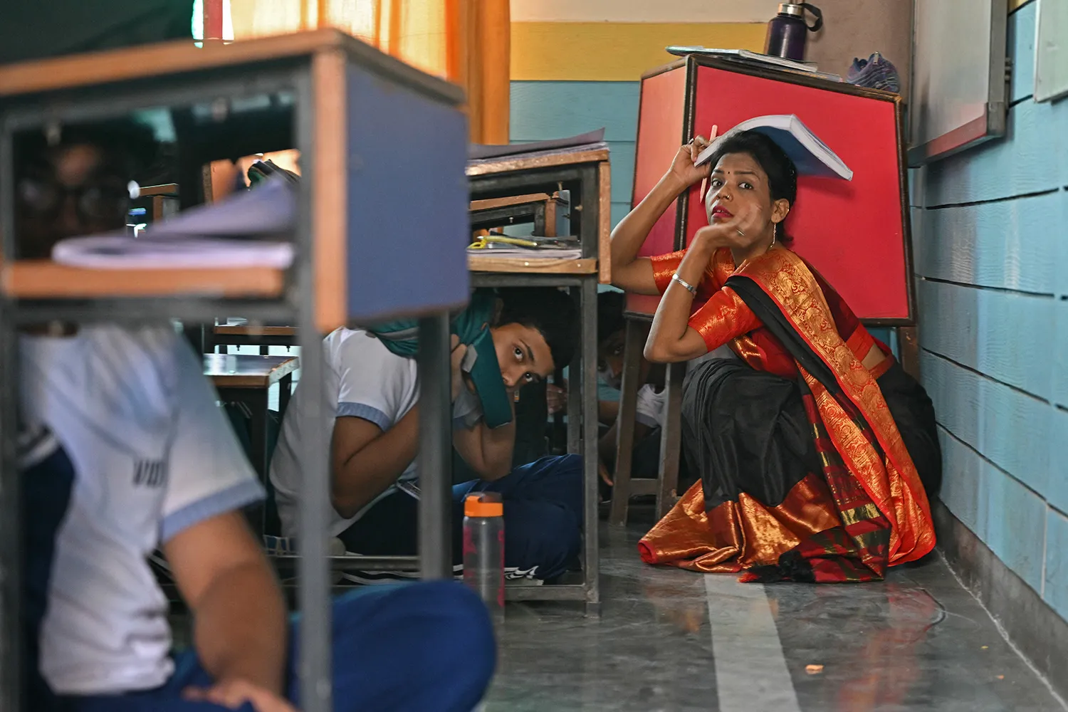 A kneeling woman looks at the camera with a notebook over her head as children look out from under desks.