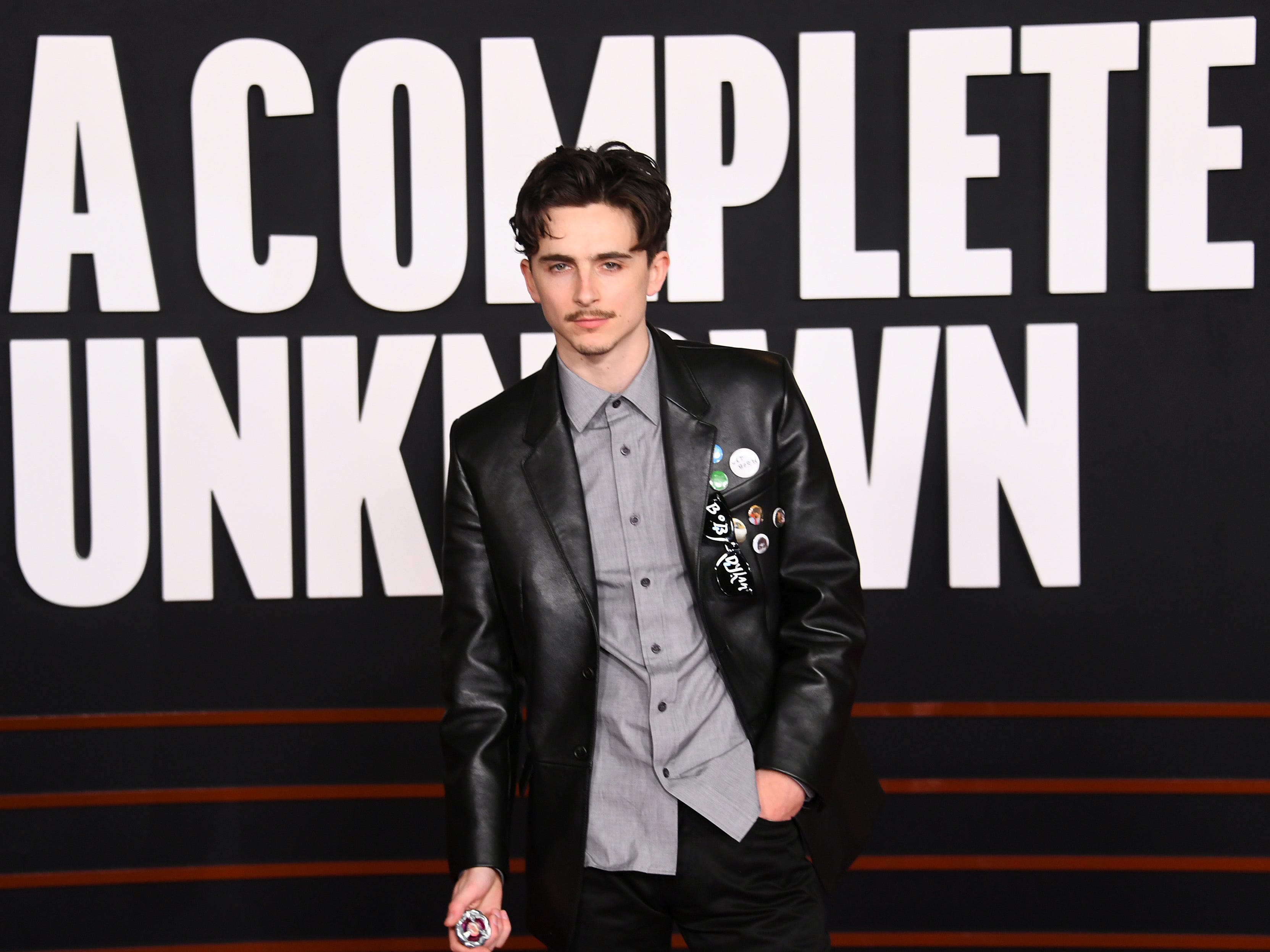 timothee chalamet at the los angeles premiere of a complete unknown, wearing an untucked grey button up shirt with a leather jacket, adorned with pins. he's standing in front of text reading