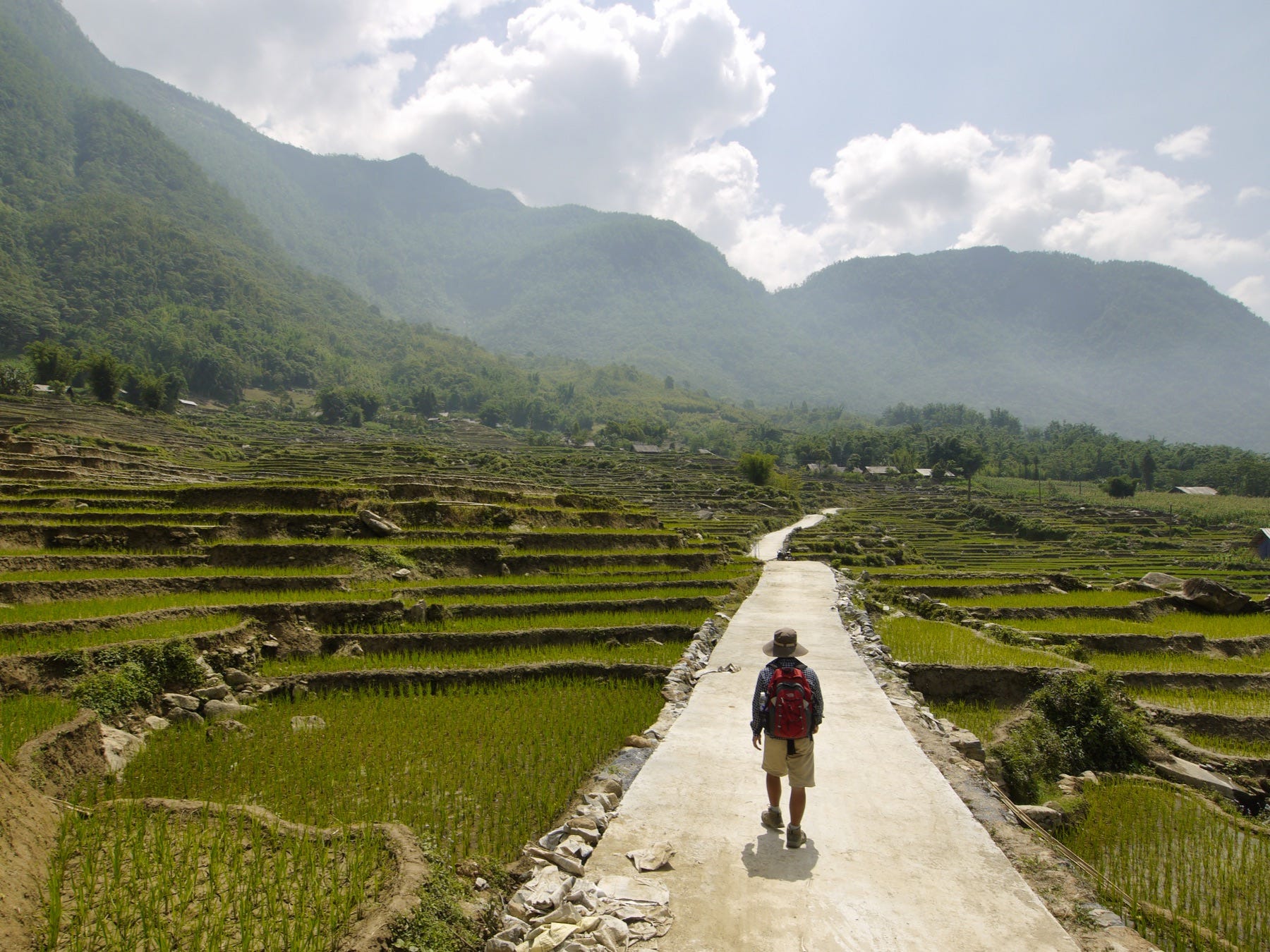 A man walking wearing a green hat and a backpack on a path between rice fields in Southeast Asia.