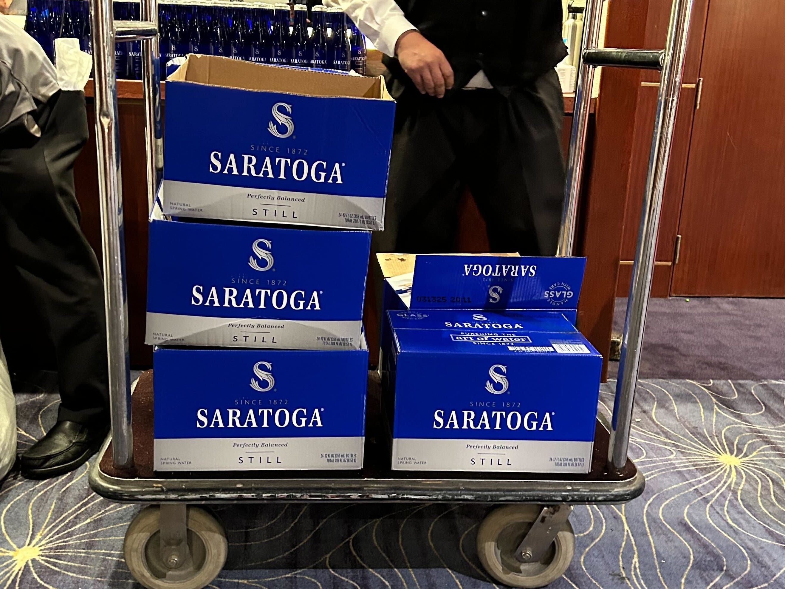 A hotel staff member moving boxes of Saratoga water on a luggage trolley.