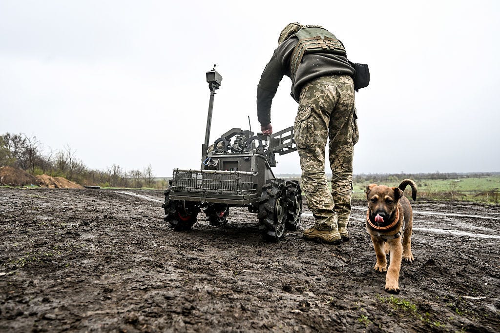 A Ukrainian soldier tinkers with a UGV on muddy ground. A puppy stands nearby.