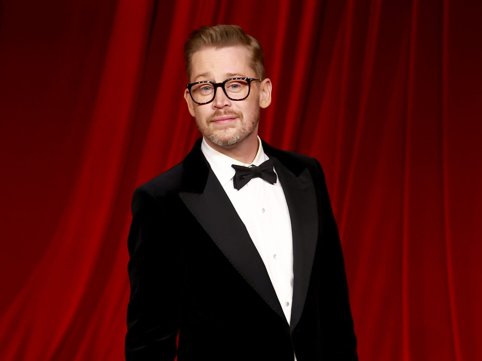 A man with short blond hair wears a black tuxedo with a white shirt and large black glasses. He has a short blond beard with gray in it. He's stood against a backdrop of red curtains.