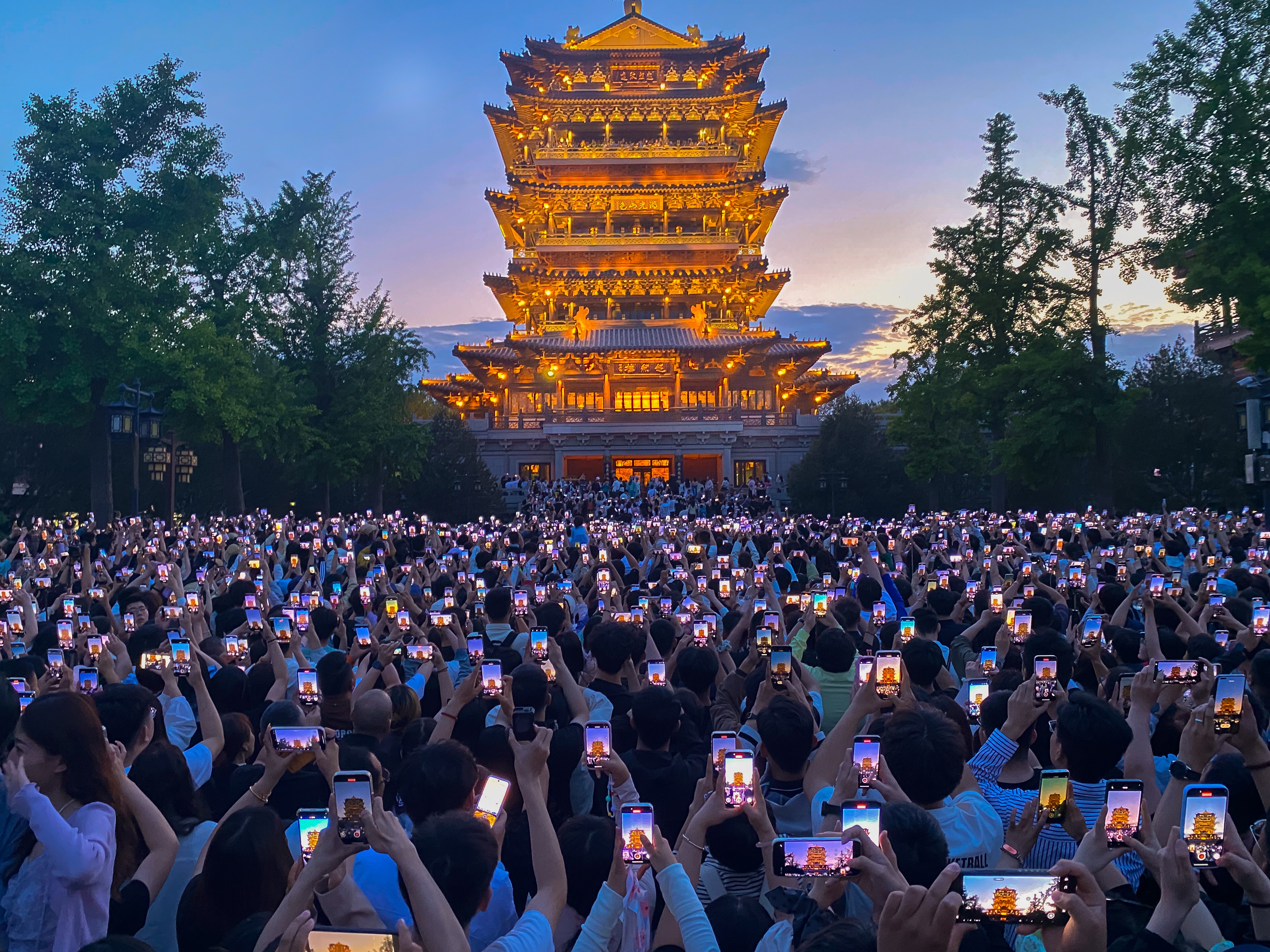 Hundreds of people stand together, holding their phones up to take photographs of an illuminated tower.