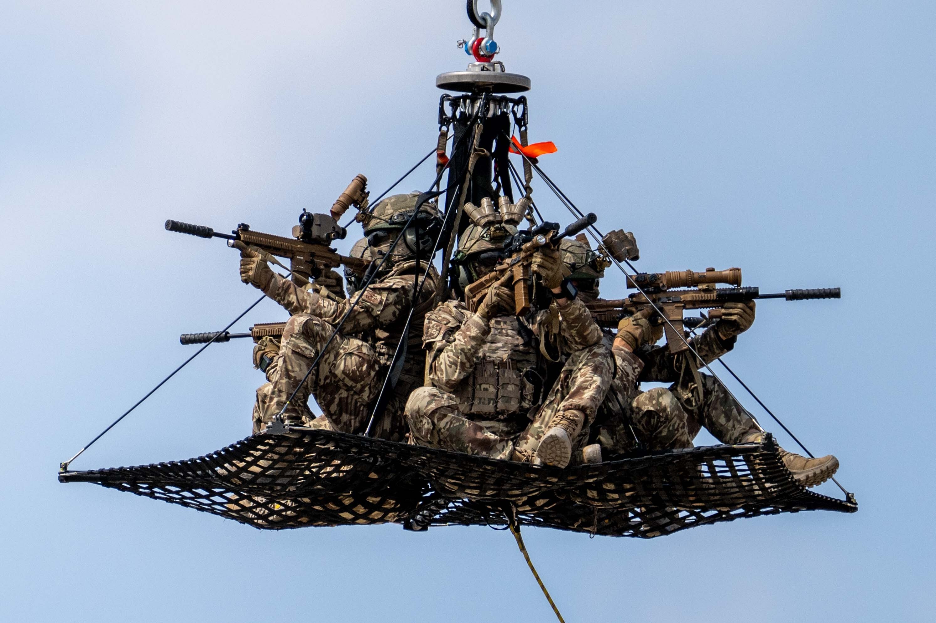 Several soldiers in camouflage gear hold weapons while being lifted in a netted container.