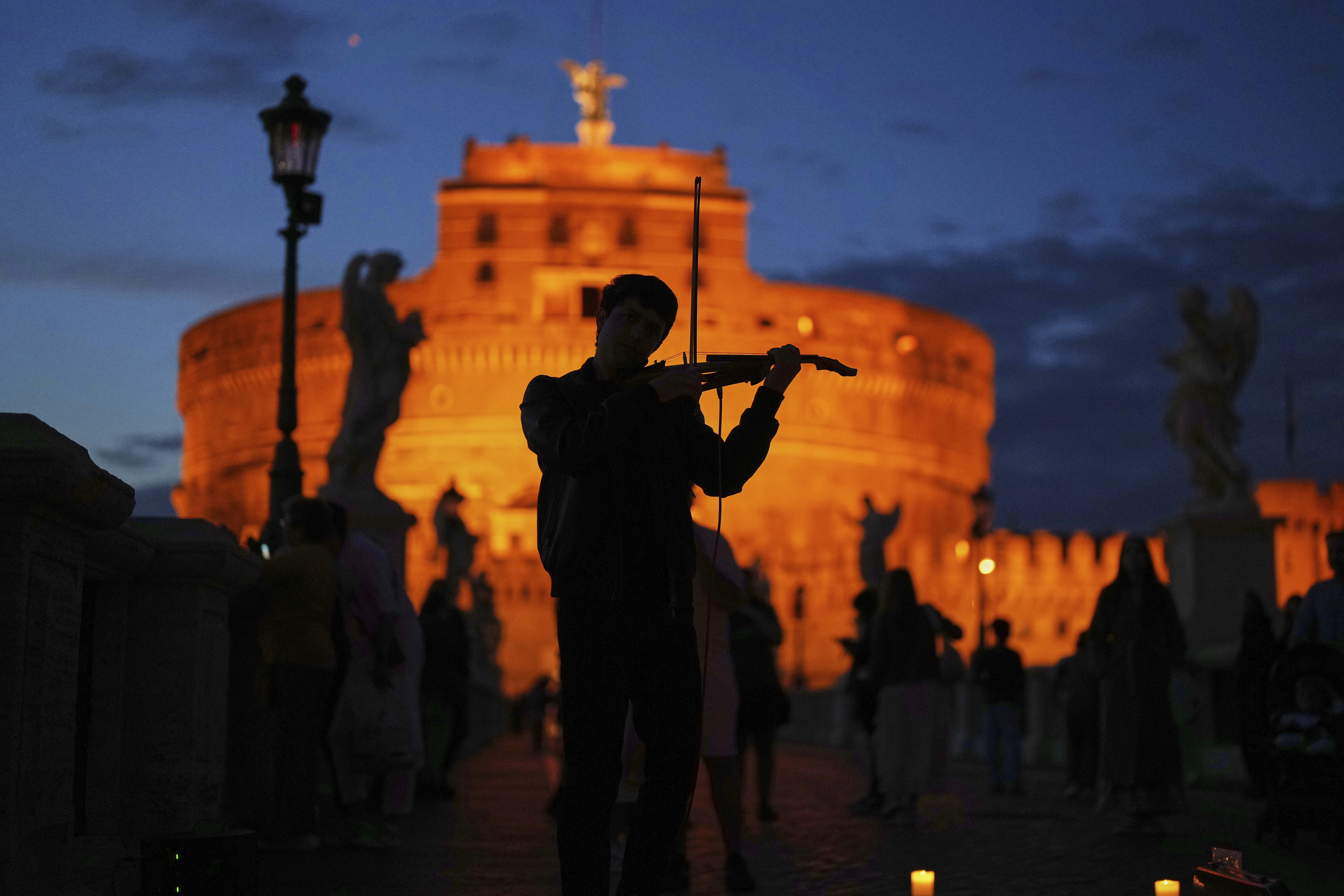 A person plays a violin, seen in silhouette in front of a building lit up by a sunset.