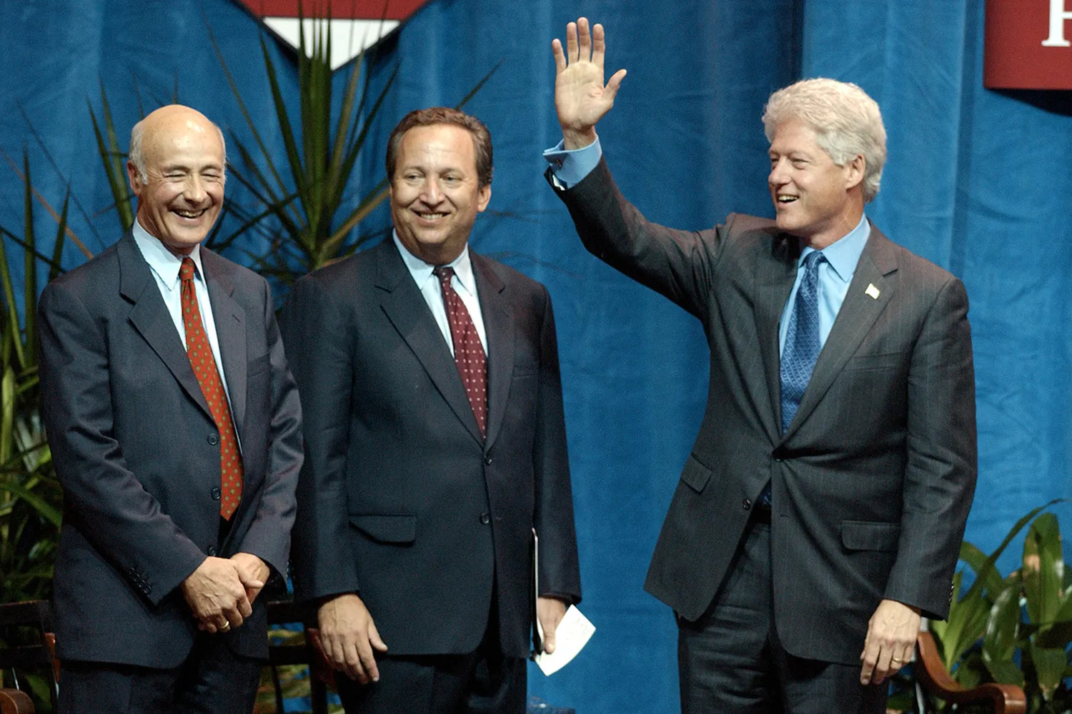 Clinton at right waves as he stands next to Nye and Summers on a stage.