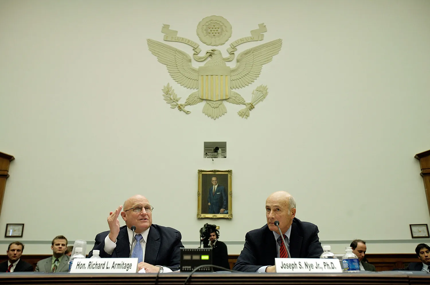 The two men sit a a table with microphones and placards bearing their names. Above them is U.S. eagle seal on the wall.