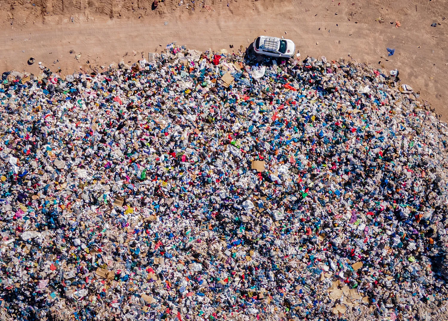 An aerial view shows a large colorful pile of clothese with a small car at the top passing buy in a dirt road.