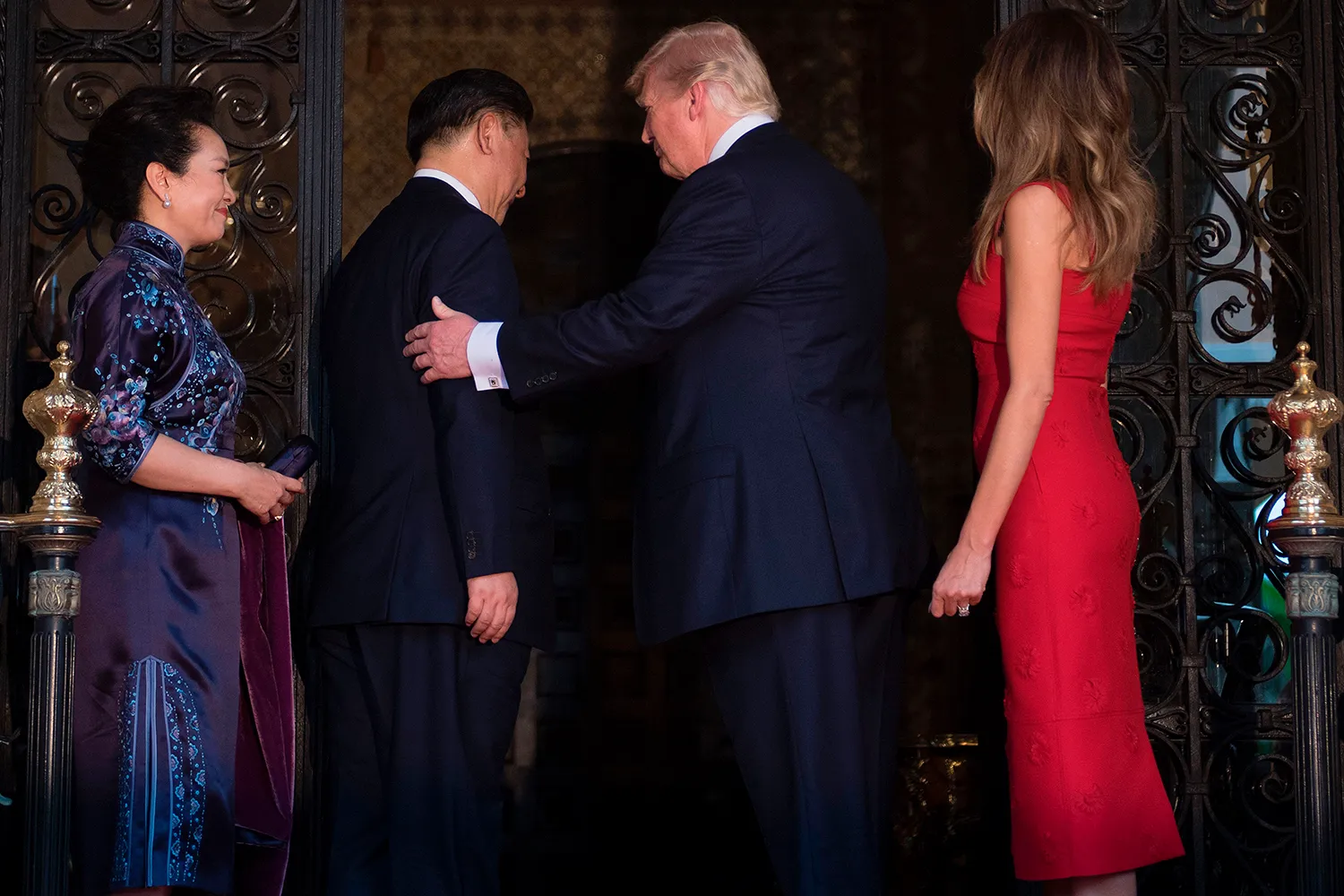 Donald Trump welcomes Xi Jinping with a pat on the shoulder as they stand in a doorway, facing away from the camera. Their wives stand on either side of them in formal, calf-length evening gowns.