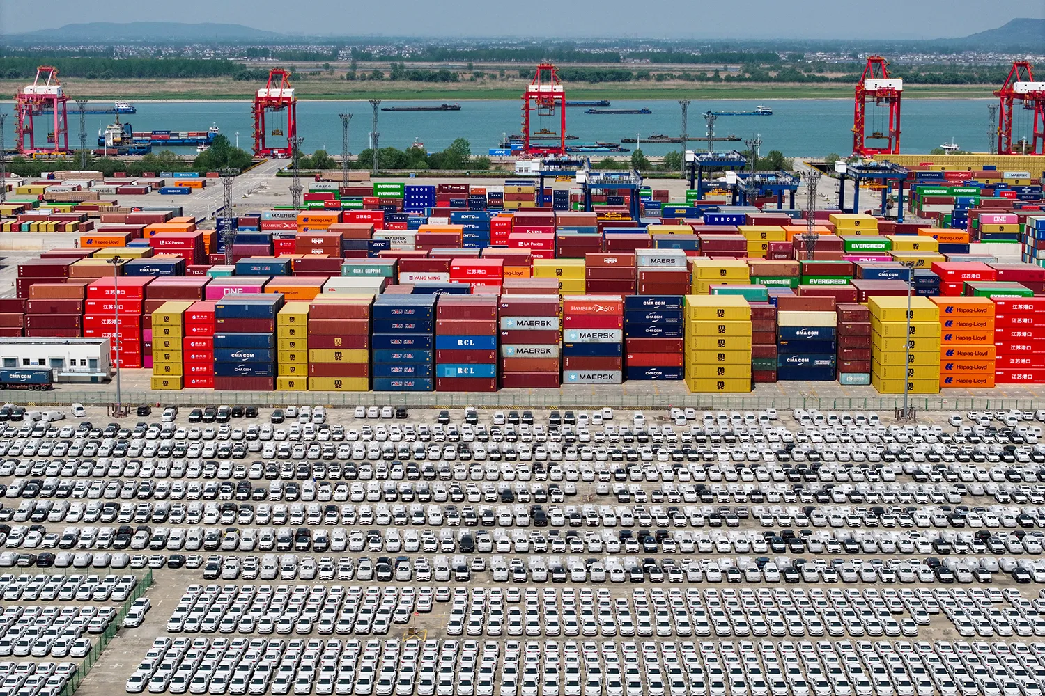Thousands of cars are seen from high above in an aerial photo, lined up in nearly perfectly neat rows in a parking lot in front of a port yard with colorful shipping containers stacked in piles. The water is visible beyond that, a few massive ships floating nearby.