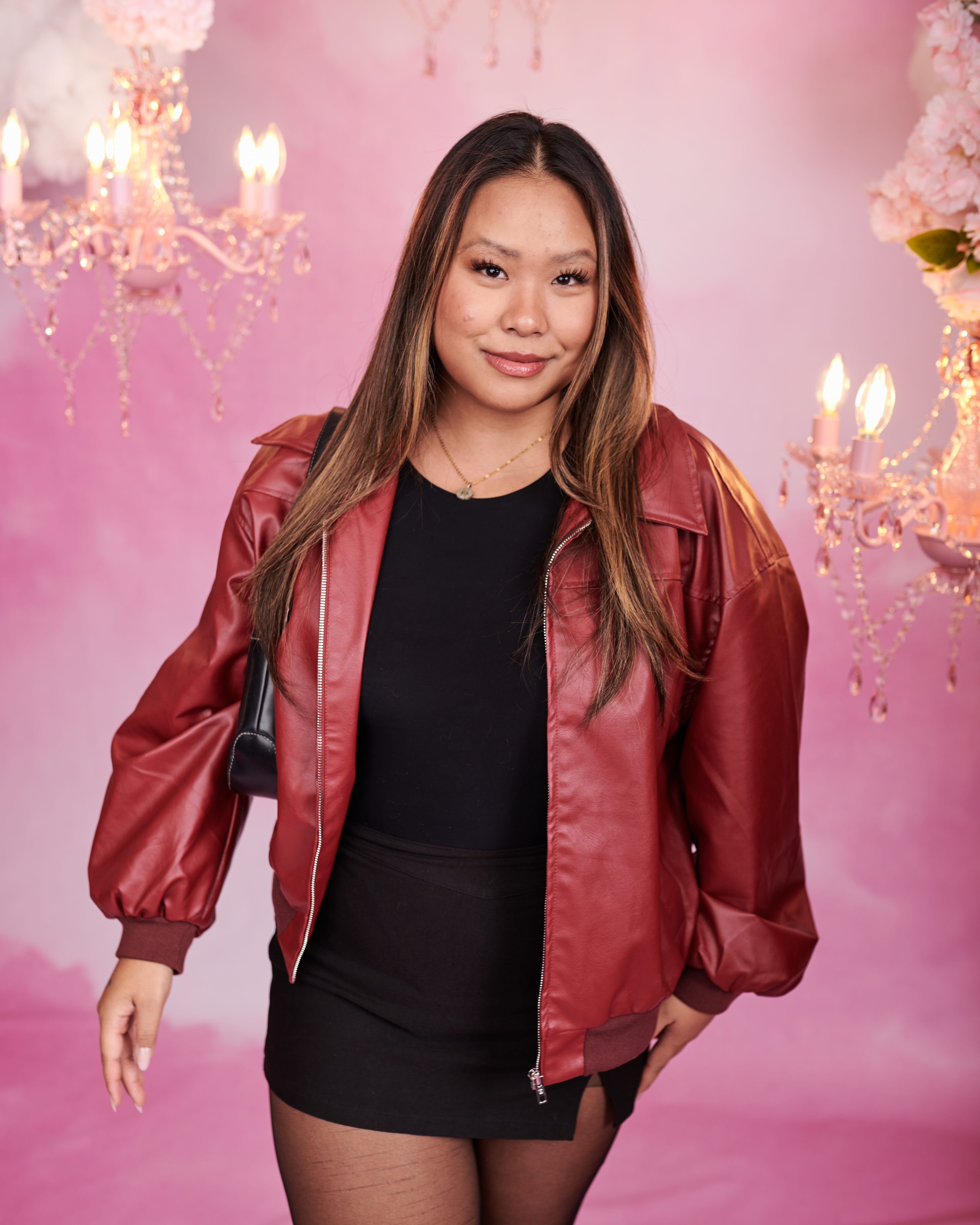 A photo of Bridgette Vong in a red leather jacket with chandeliers and a pink background.