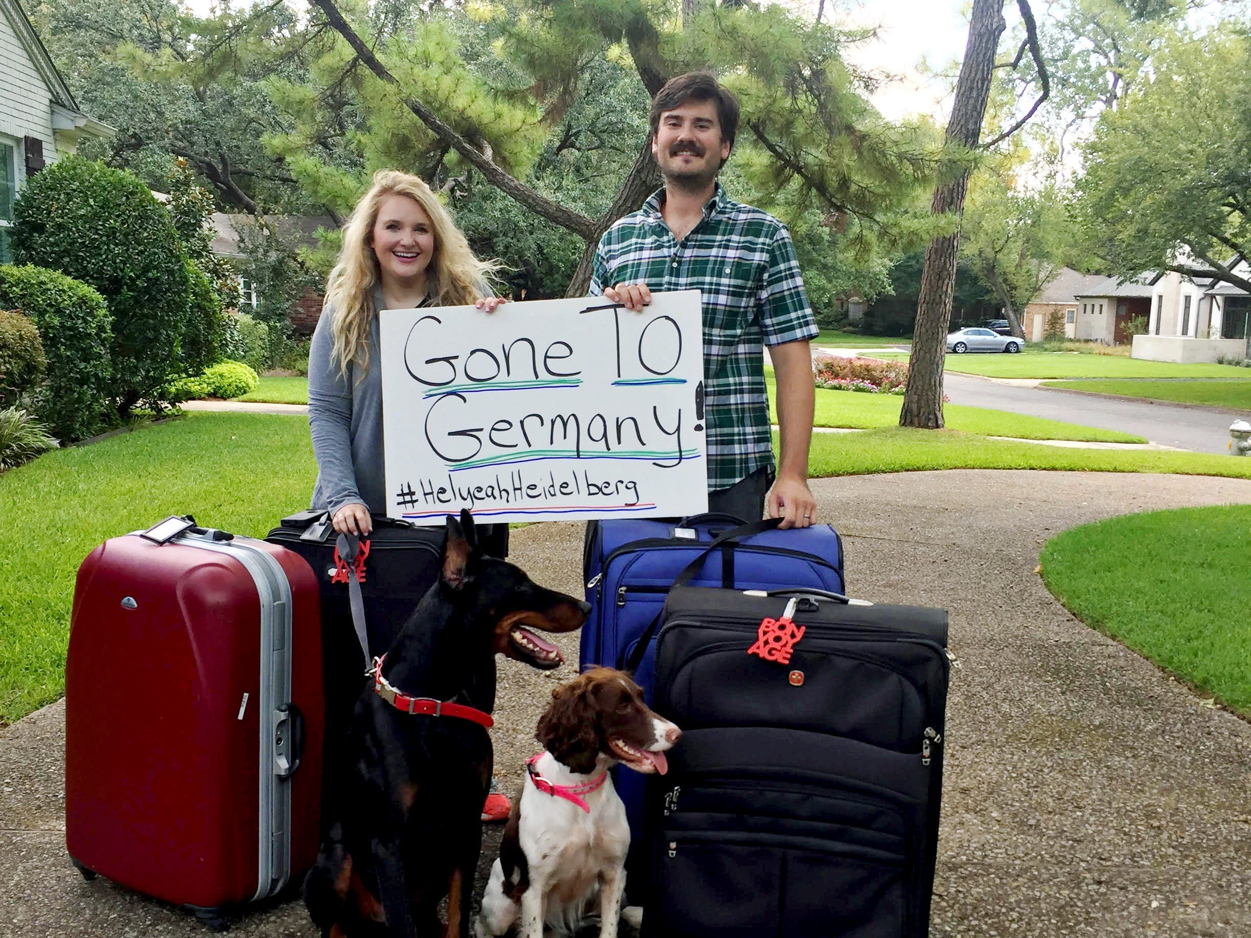 A woman and a husband posing in a large driveway while holding a handwritten sign that says "Gone to Germany! #HelyeahHeidelberg." At their feet are four large suitcases and two dogs.