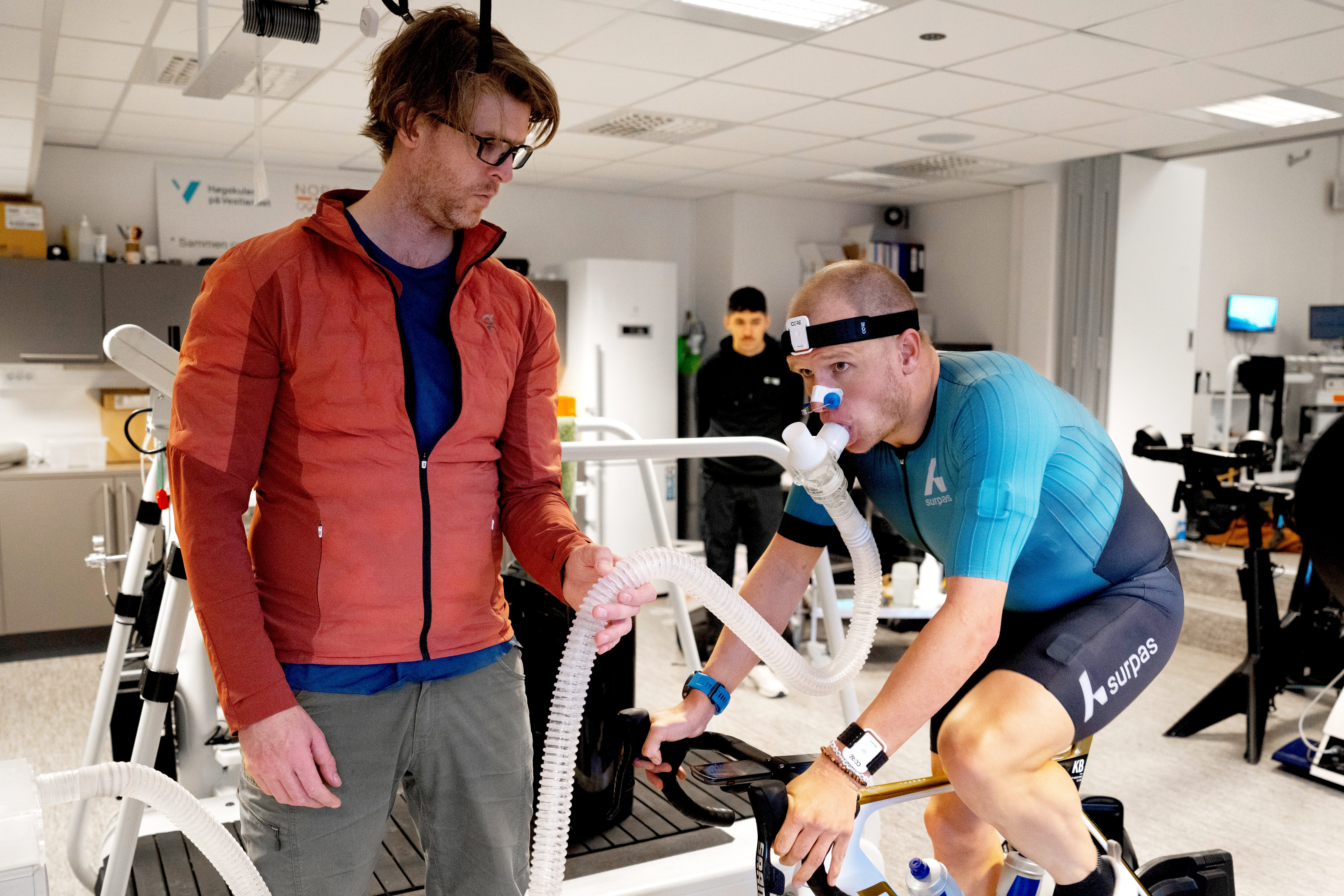 A man on an exercise bike is performing an endurance test in a lab as his coach looks on.