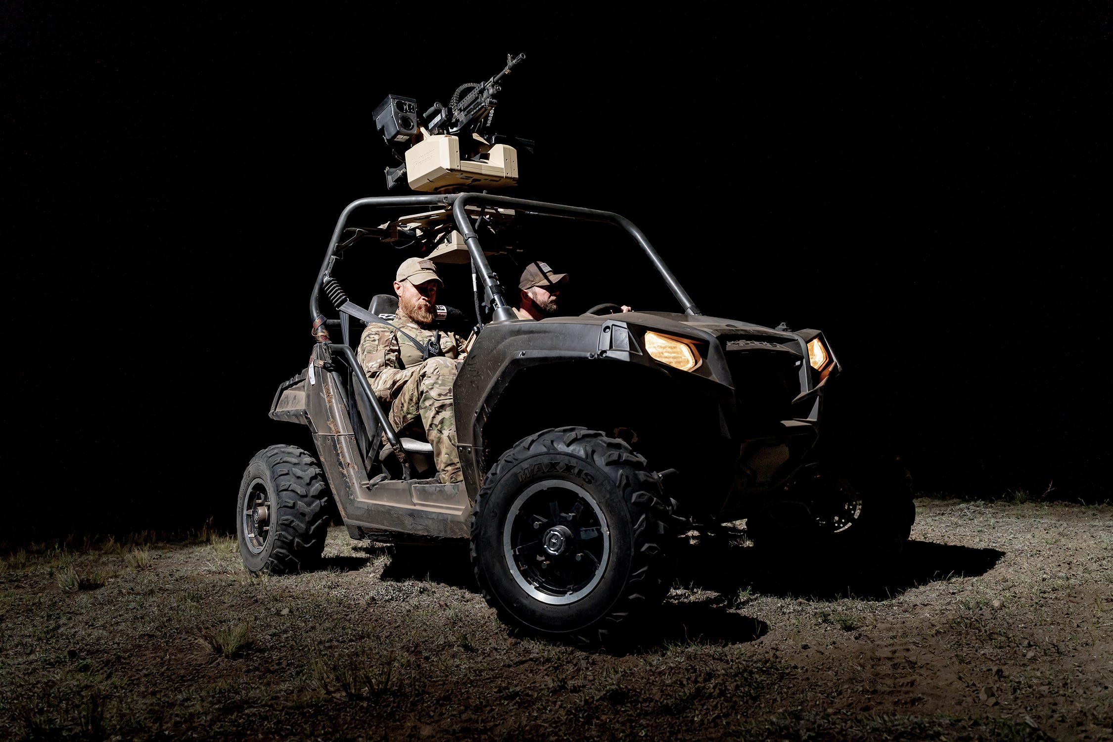 Two men sit in a vehicle with an automated machine gun on top of it at night.