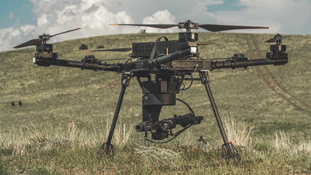 An AimLock system on an uncrewed aerial vehicle sitting in a field with a hill and cloudy sky in the background.