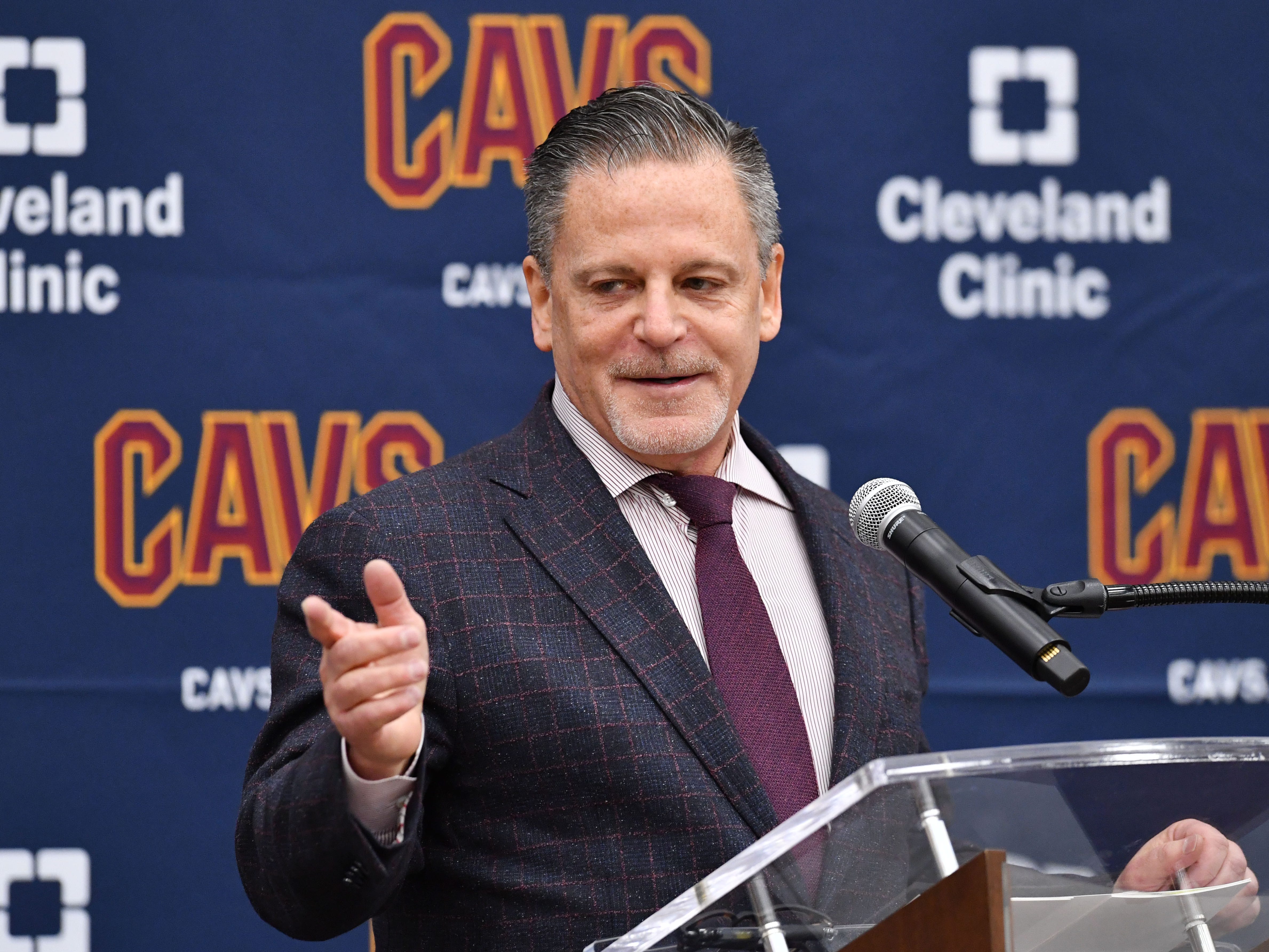 Dan Gilbert behind a microphone with a blue Cleveland Cavaliers backdrop with Cleveland Clinic logos on it