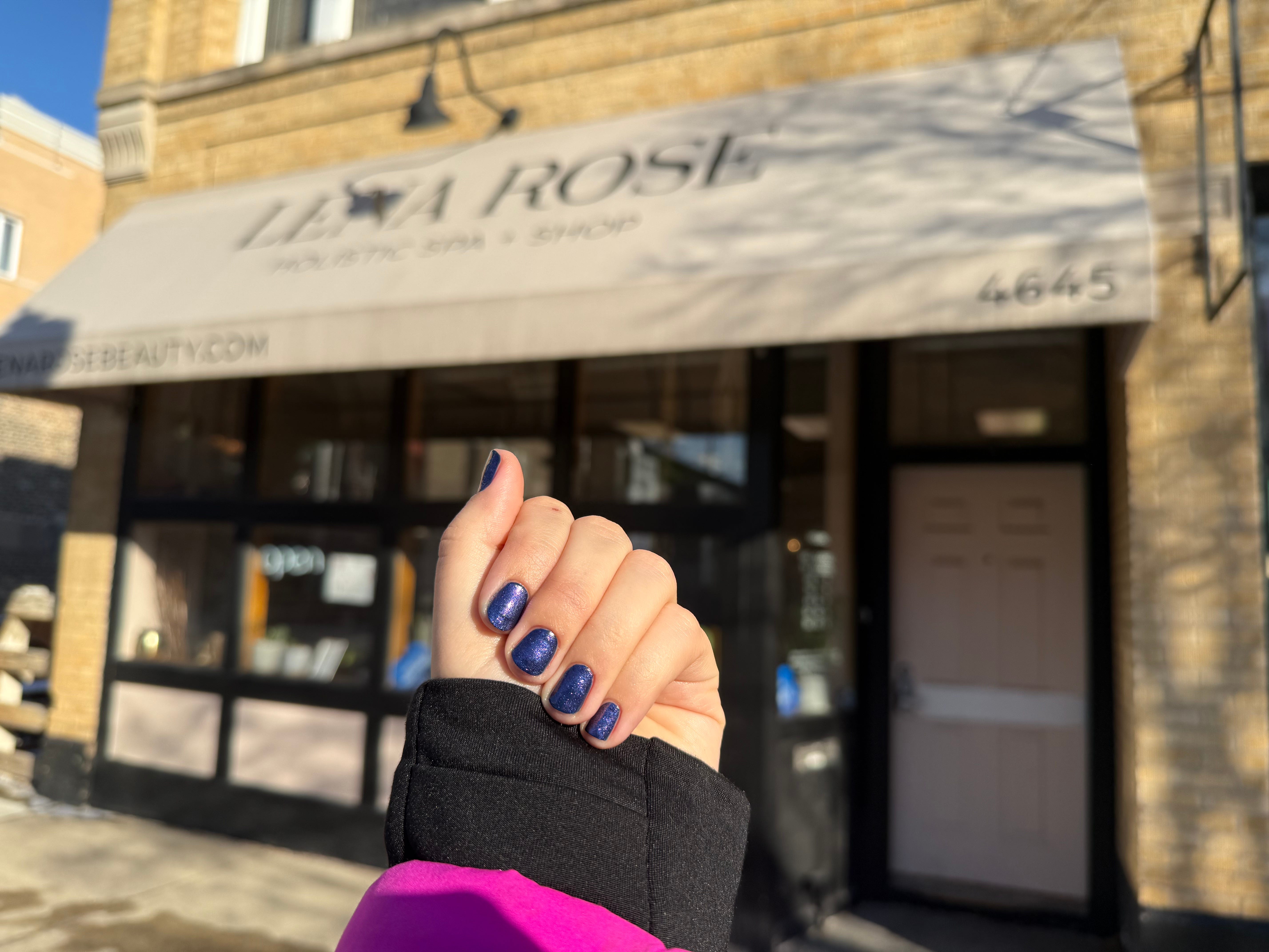Gia shows off her nails with sparkly purple polish in front of the exterior of a nail salon.