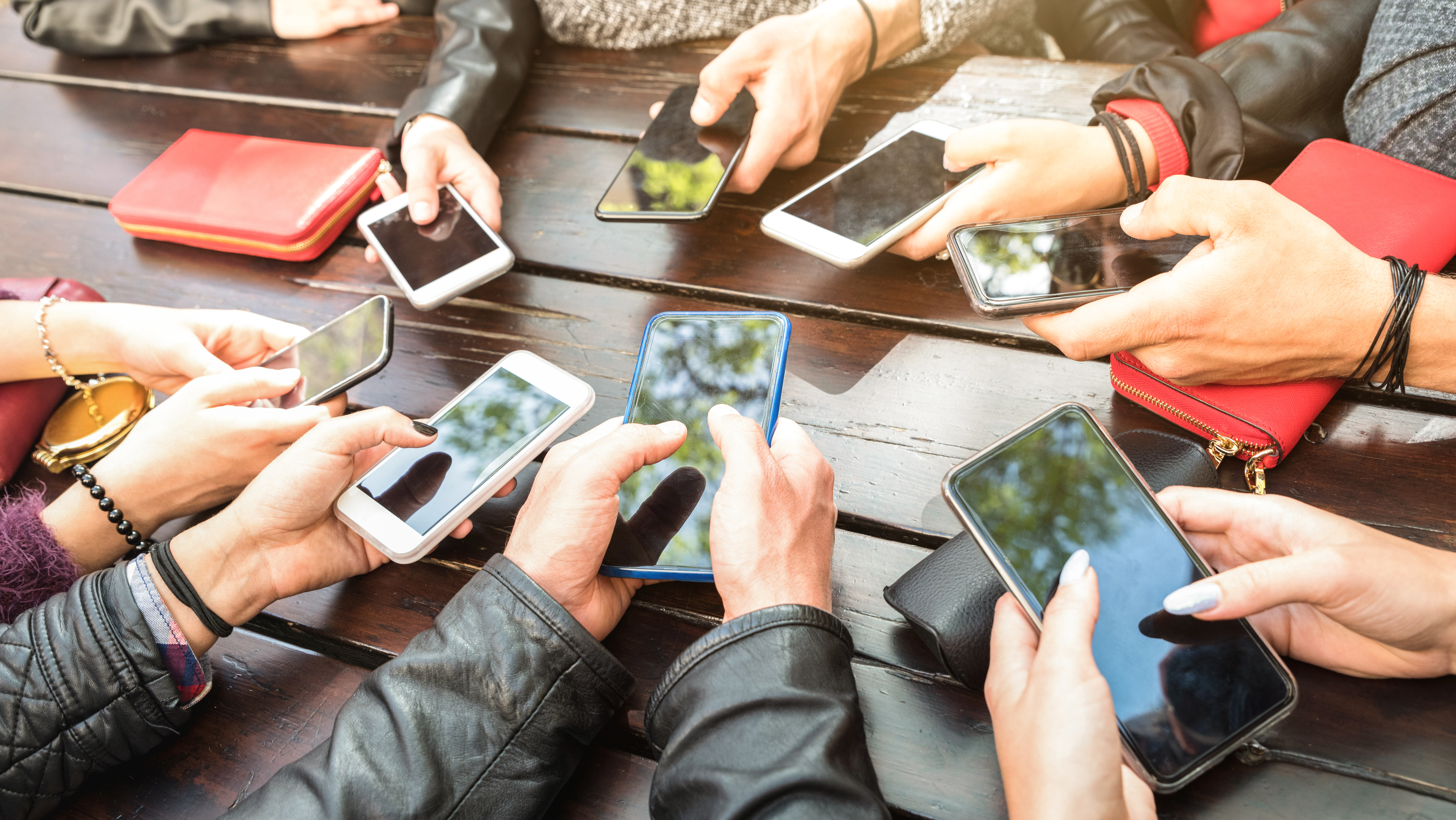 Young people sitting at table, only their hands seen in circle all on phones outstretched 