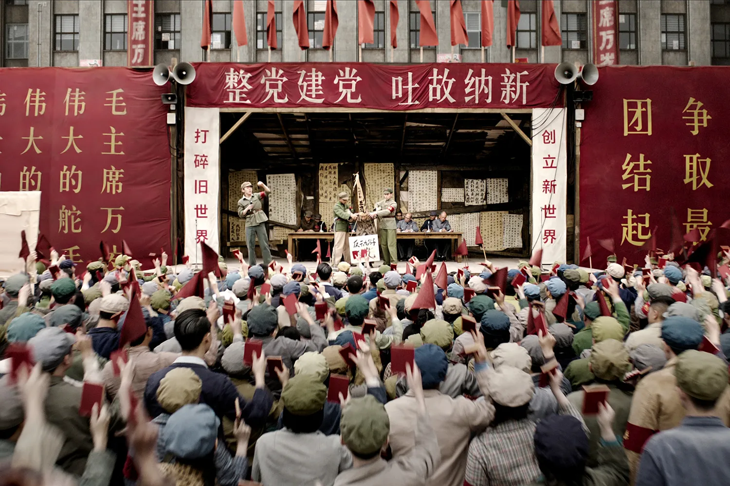 A crowd of people holding Mao's Little Red book aloft cheer as a man is tortured on a stage bearing words in Chinese and red flags.