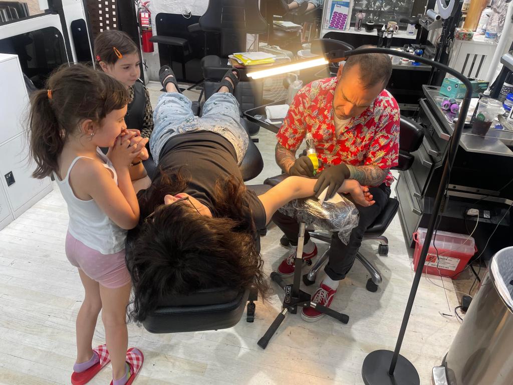 Two young kids watch their mom get tattooed on Mother's Day at Live By The Sword in Williamsburg, Brooklyn.