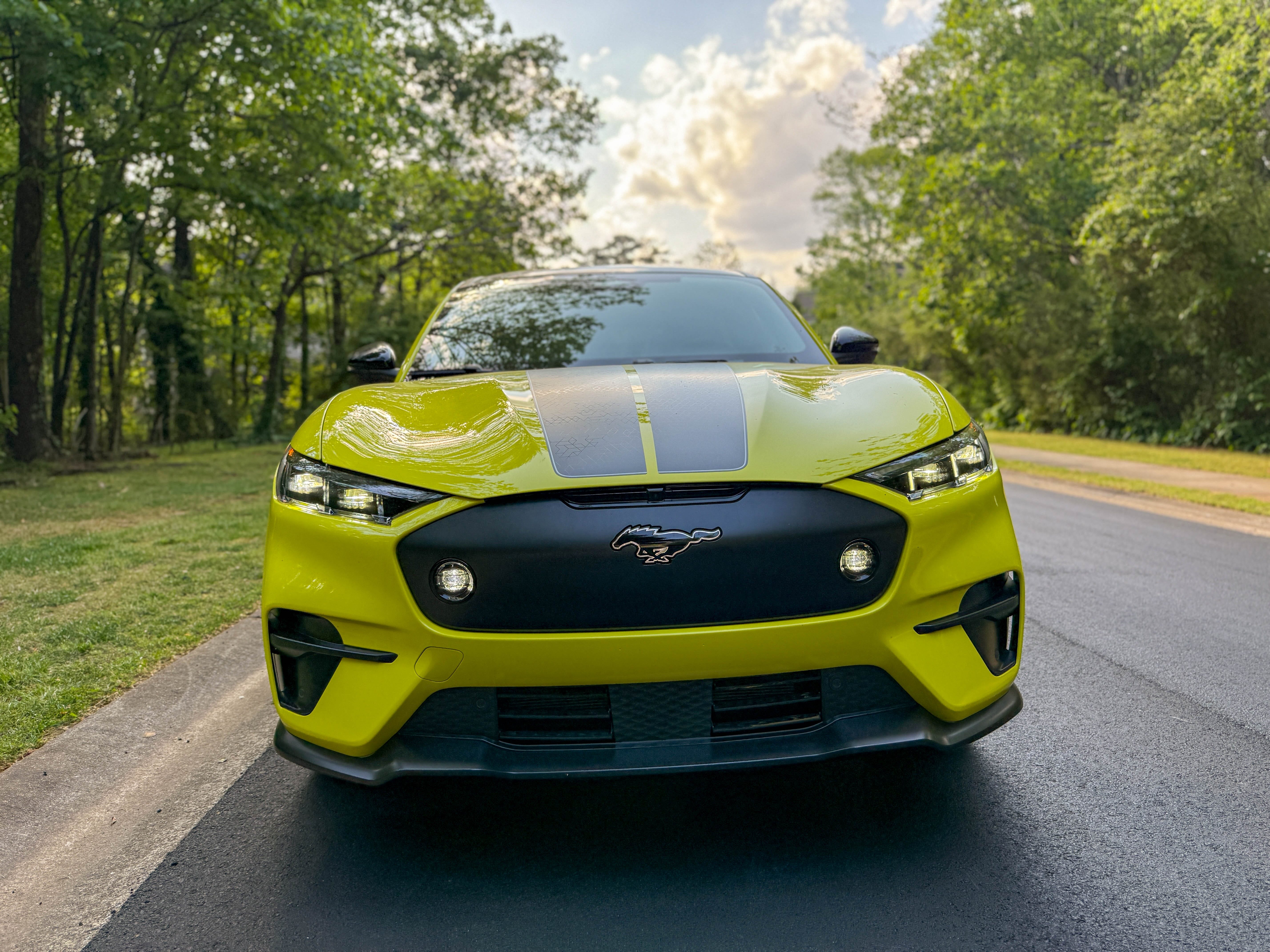 The front of a yellow 2025 Ford Mustang Mach-E Rally EV SUV parked by the woods.