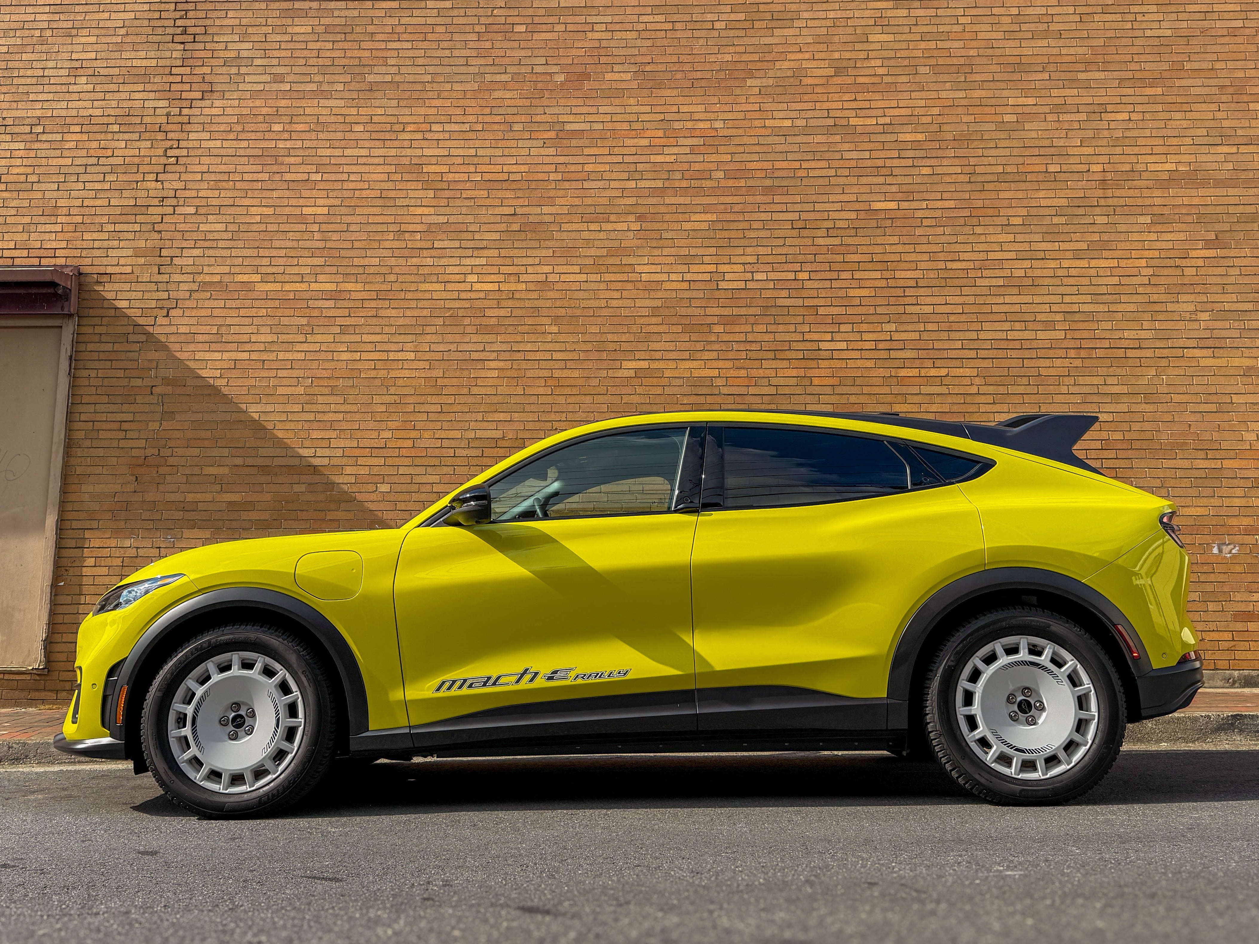 The left side of a yellow 2025 Ford Mustang Mach-E Rally EV SUV parked on the street in front of a wall.