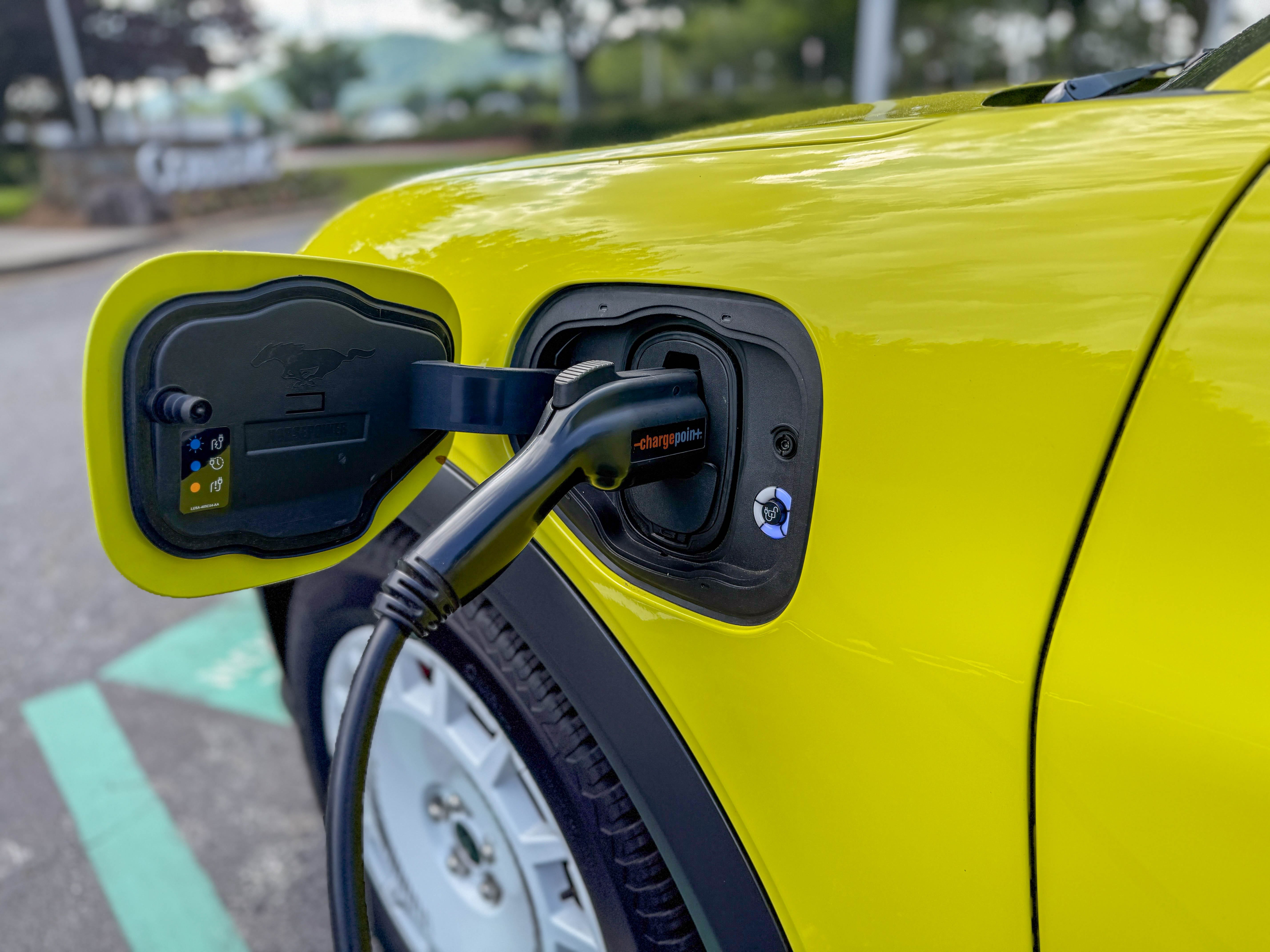 The charging socket on the left front fender of a yellow 2025 Ford Mustang Mach-E Rally EV SUV with a ChargePoint charger plugged in.