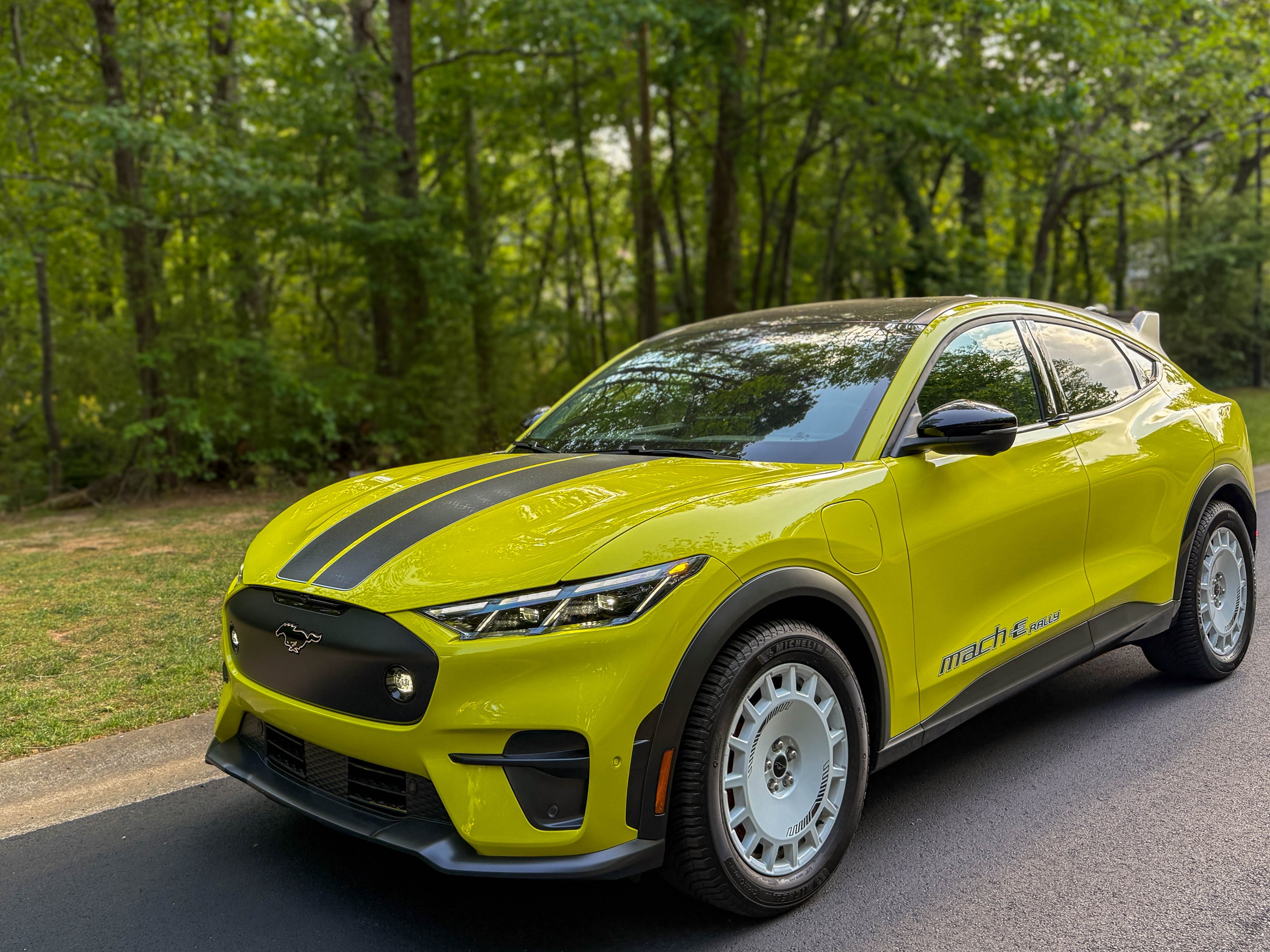 The left front of a yellow 2025 Ford Mustang Mach-E Rally EV SUV parked by the woods.