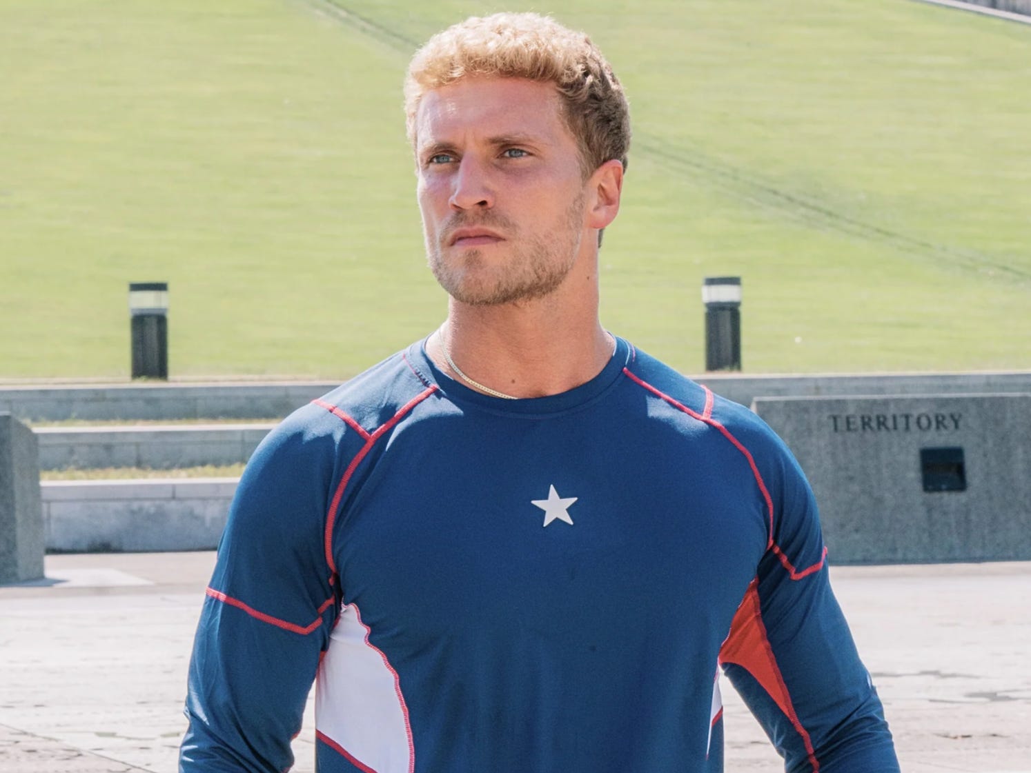 A man with blond hair and short stubble is standing in a concrete square park; there is grass behind him. He's wearing a blue long-sleeve top with red outlines around the shoulders, down his ribs, and down his forearms. There is a white star in the center of his chest. There is a white panel on his ribs.