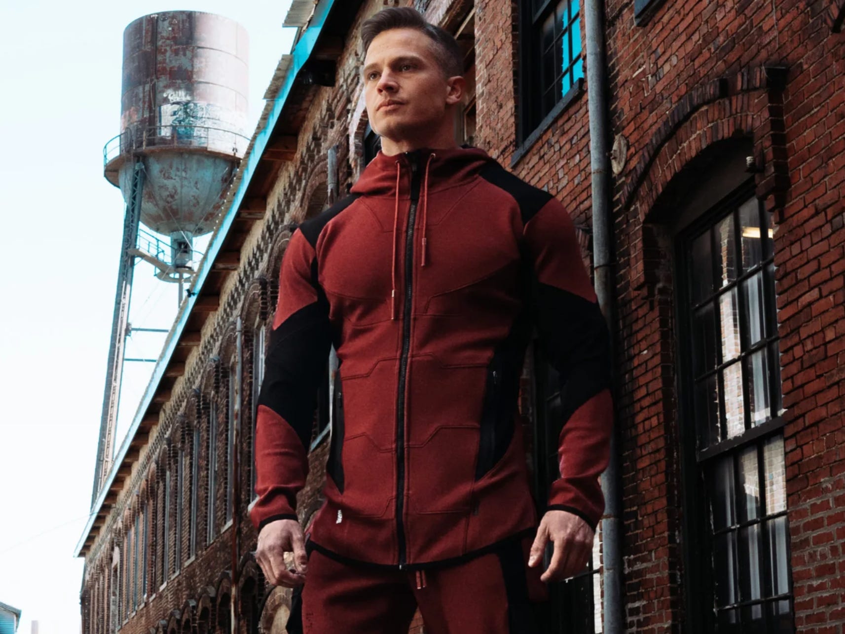 A man with short brown hair stood in front of a red-brick building. He's wearing a red and black tracksuit with detailed stitching to make it look armored.