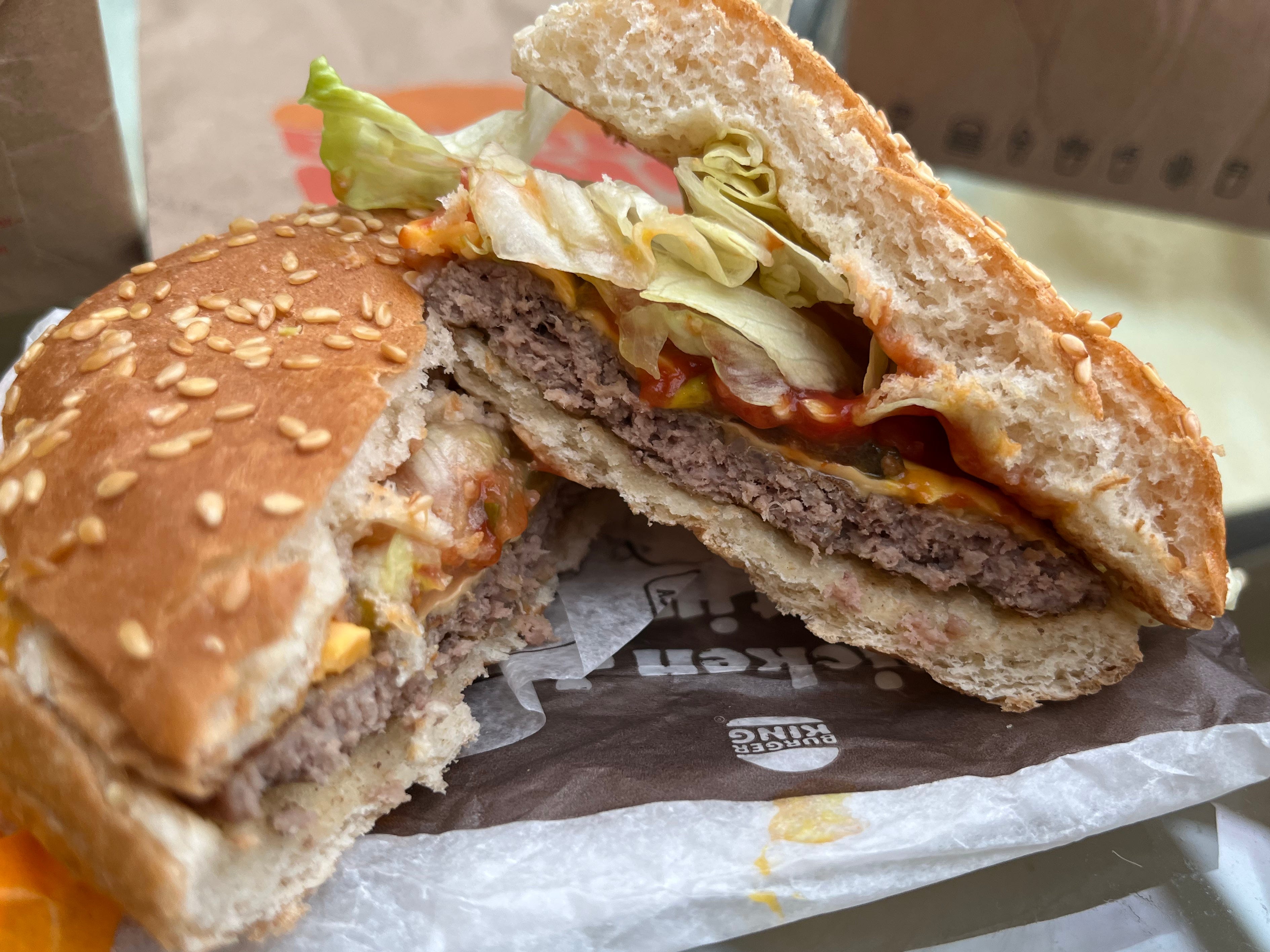 Two halves of a cheeseburger with lettuce, tomato, pickles, and a sesame seed bun stacked on top of each other.