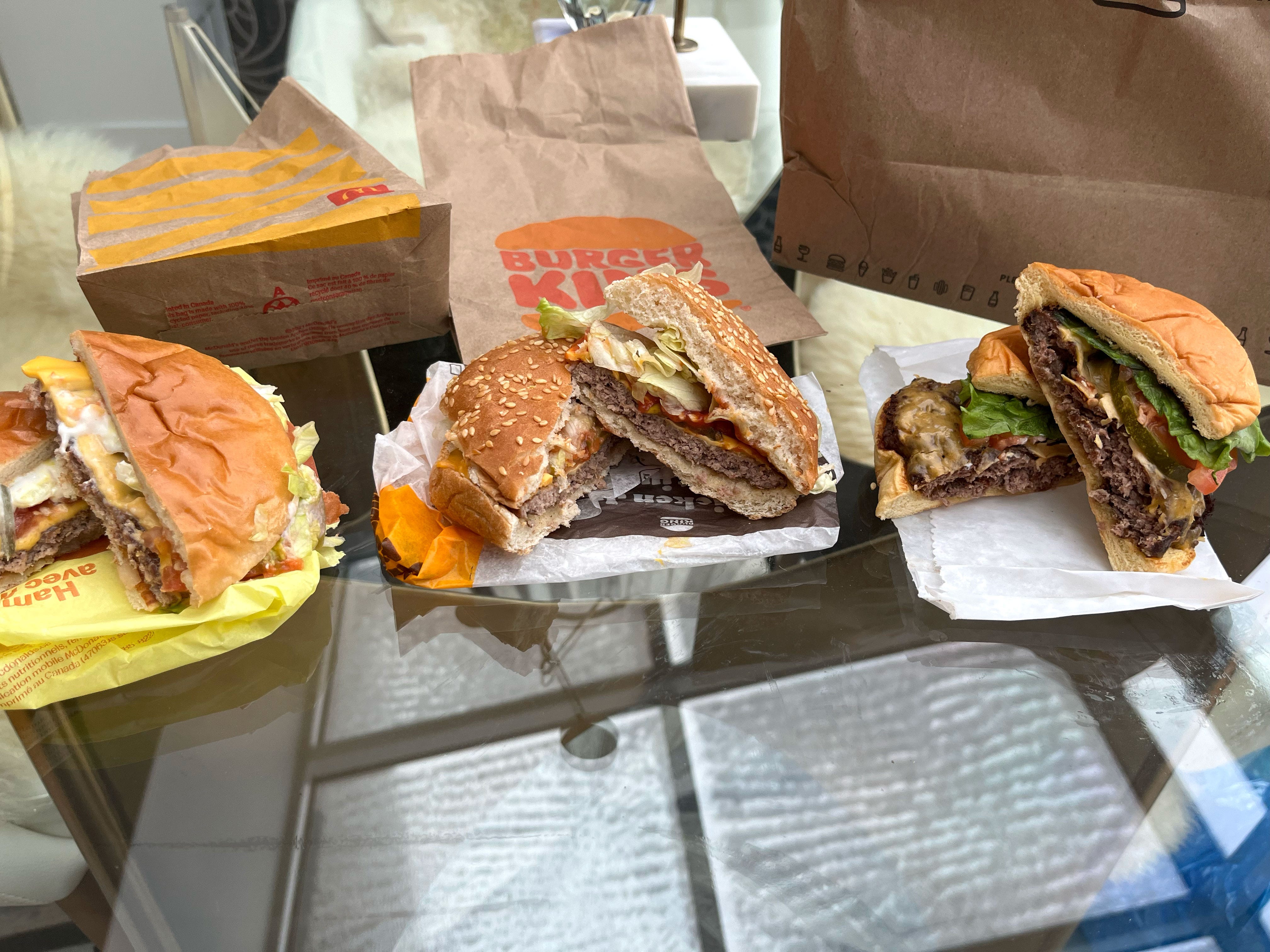 Three cheeseburgers cut in half next to bags from McDonald's, Burger King, and Shake Shack.