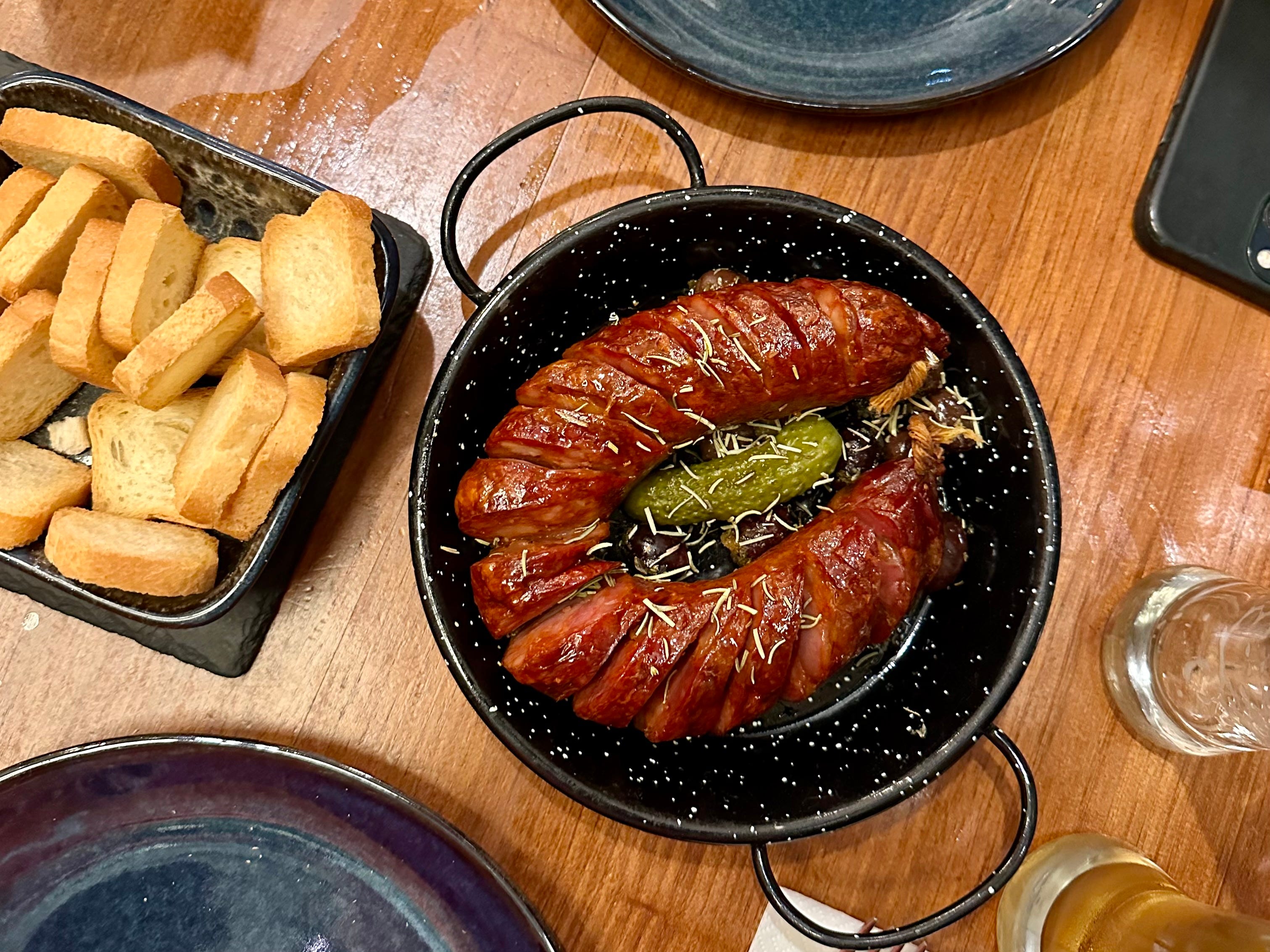 A sausage ring cut into pieces with a pickle in the middle in a black pot. Off to the side, there is a black dish filled with toasted slices of bread.