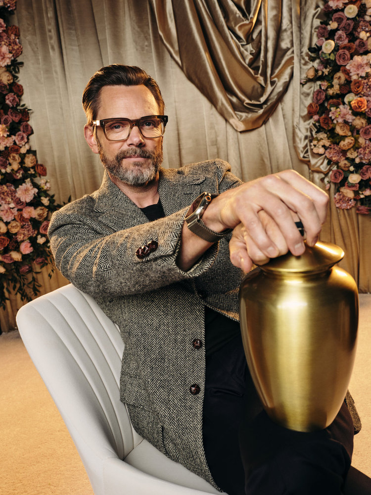 Joel McHale in a tweed jacket, black t-shirt and black pants.