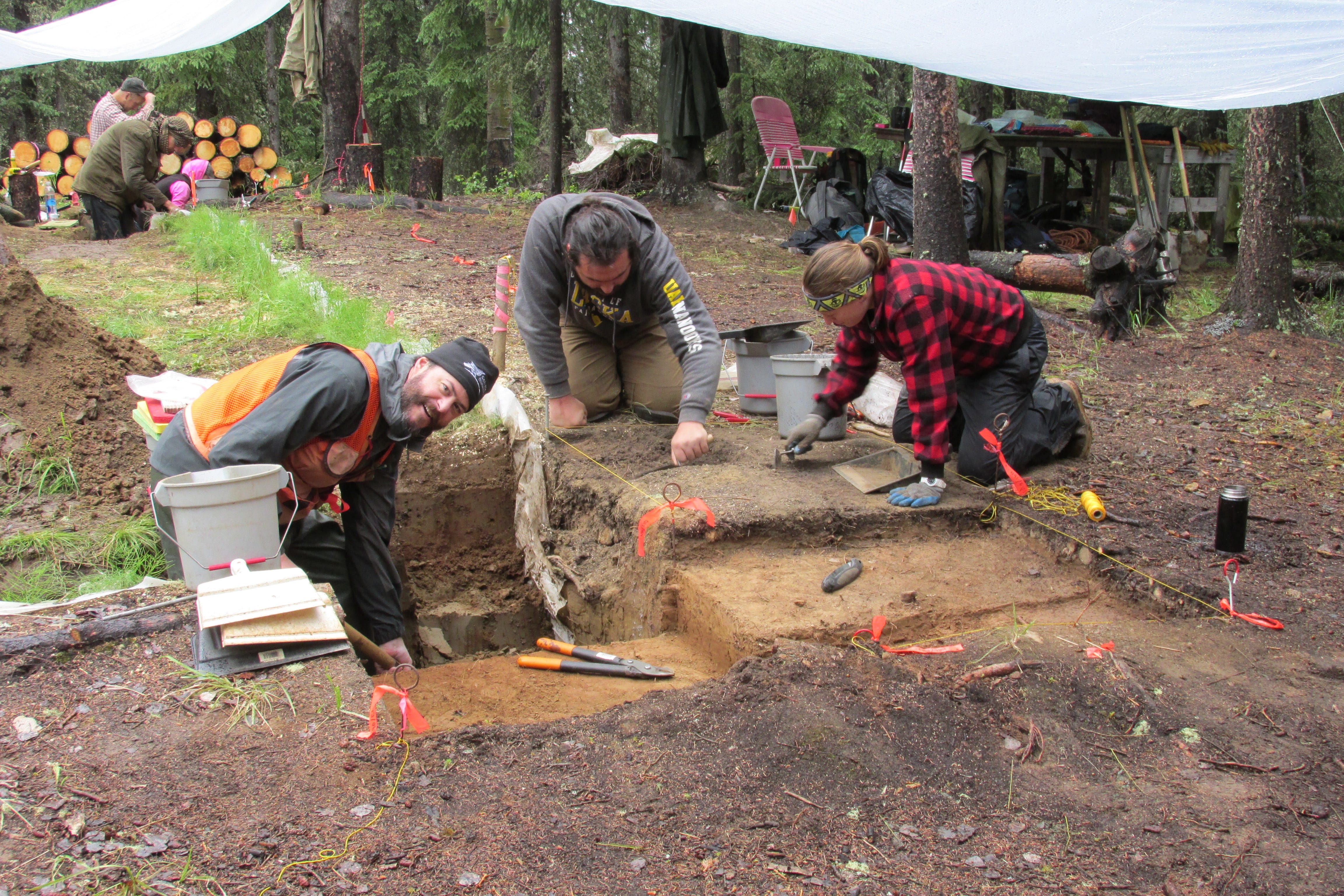 Three people crouch near a rectangular hole in the ground surrounded by excavating tools