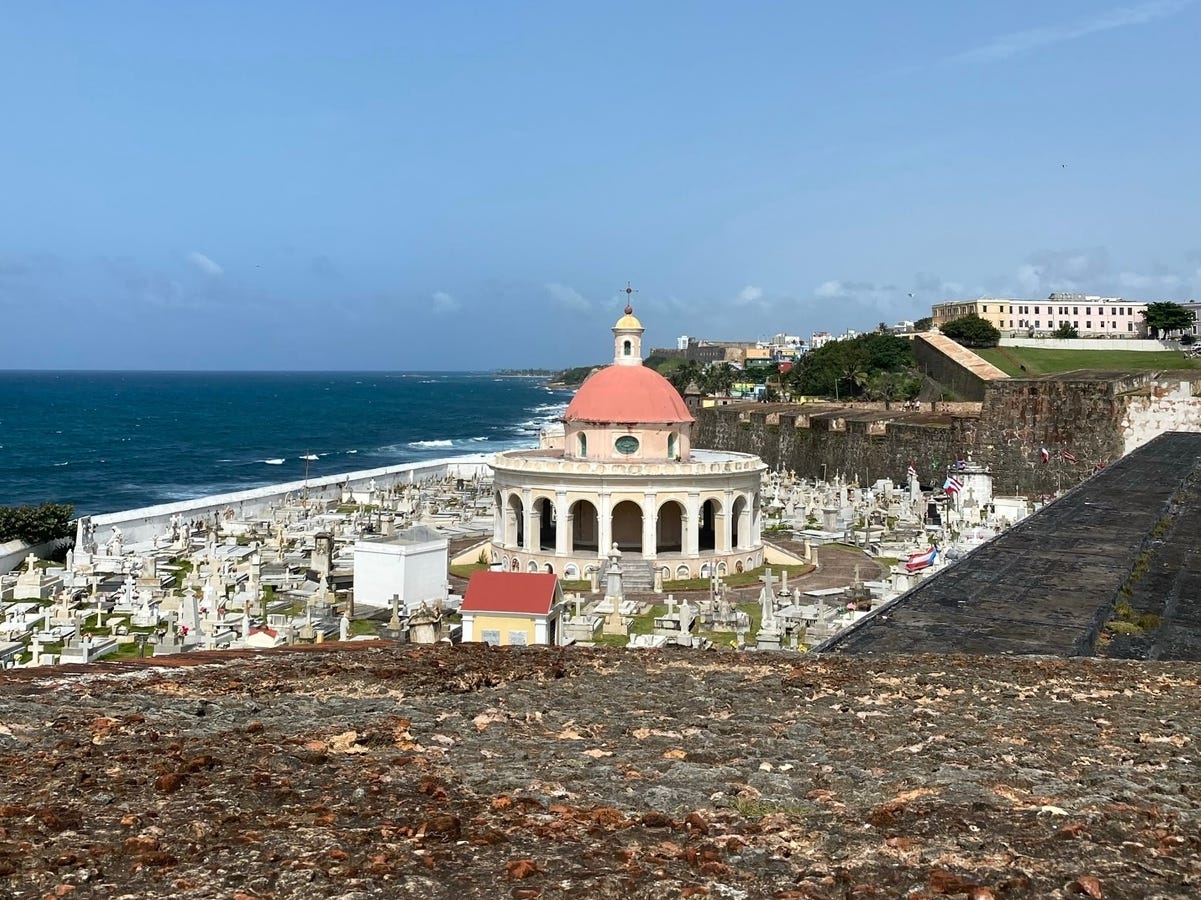The view from above of a circular building with a red domed roof, surrounded by gravestones, and nestled next to the ocean.