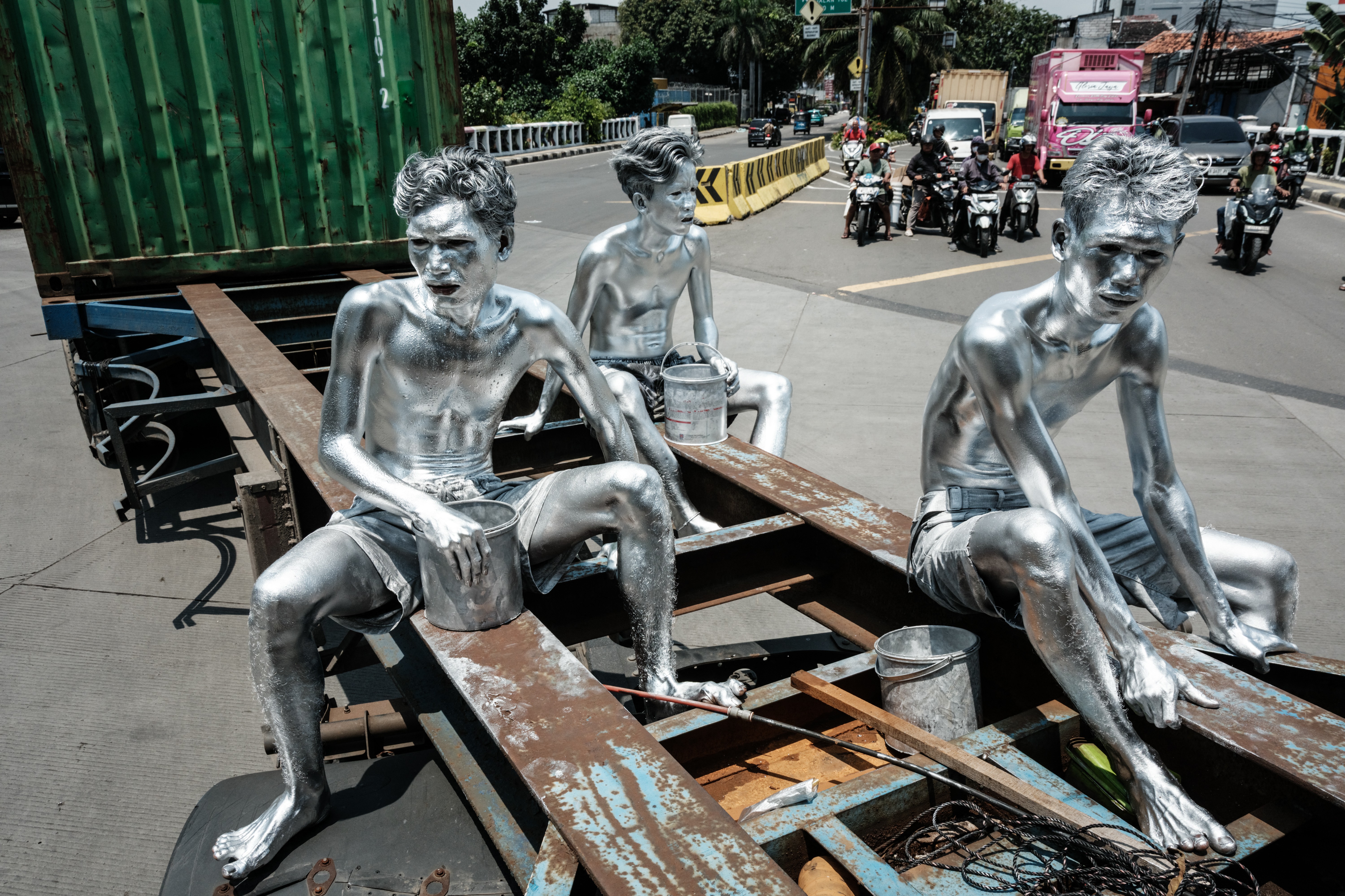 Three men covered in silver paint ride on the back of a container truck.